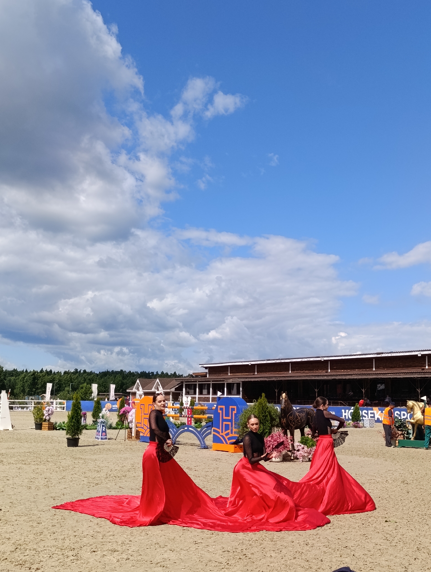 Two women in vibrant red flamenco dresses and black tops are posing on a sand-covered ground, likely an equestrian arena. The woman on the left is seated, holding a fan, and looking towards the camera. The woman on the right is standing, also holding a fan, and looking away from the camera. Both women have their arms gracefully positioned.
In the background, there is a wooden building with a brown roof, possibly a stable or grandstand, and a line of trees. The arena is decorated with floral arrangements, ornamental trees, and some equestrian-themed statues or props, including a golden horse statue and a dark, ornate horse sculpture. There are also various signs and banners in the background, some with blue and orange elements. The sky is bright blue with scattered white and grey clouds, suggesting it is a pleasant day, likely daytime. The overall atmosphere appears to be a cultural or sporting event, possibly with a performance element, given the elaborate costumes.
Visible text includes "HO SEK SPO" on a blue banner and what appears to be "ИВАНОВСК" partially visible on another banner. The location context is Ivanovskoe, Russia.