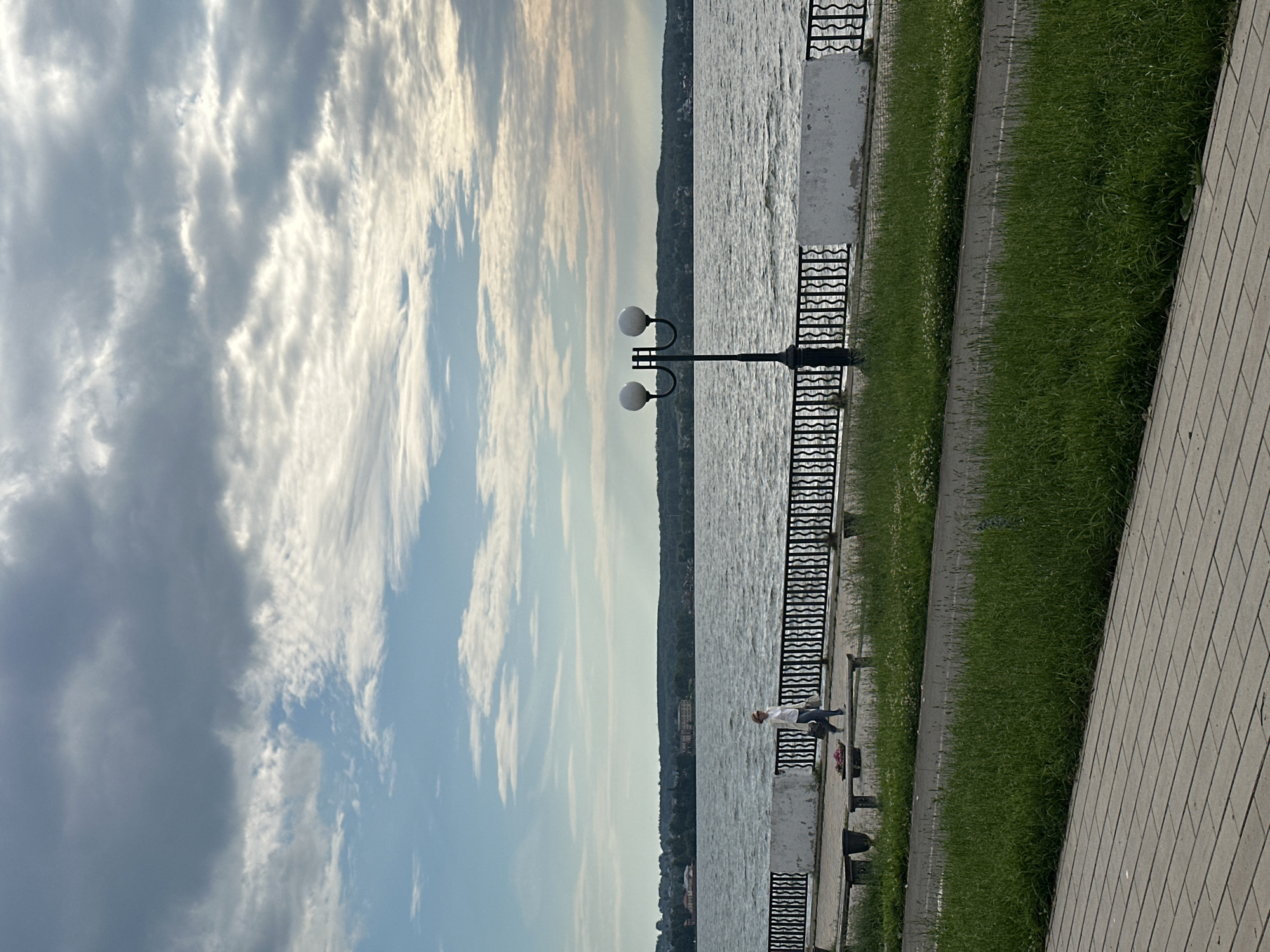 The image depicts a serene waterside promenade in Votkinsk, Russia. The scene is captured vertically, with the sky dominating the left half and the water, promenade, and grassy area on the right. The sky is filled with dramatic, wispy clouds, suggesting either early morning or late afternoon light. The water is rippling, reflecting the sky and creating a shimmering effect.
On the promenade, a single person is seated on a bench, facing the water. They are wearing a white top and dark pants. Their posture suggests relaxation or contemplation. A distinctive double-headed lamppost stands behind them, a common feature of urban promenades. A decorative black metal fence separates the promenade from the water.
Adjacent to the promenade is a strip of vibrant green grass, which then transitions to a paved walkway or road, and further to a tiled pathway. The overall impression is one of tranquility and pleasant weather, ideal for a leisurely stroll or quiet reflection by the water. There is no visible text in the image.