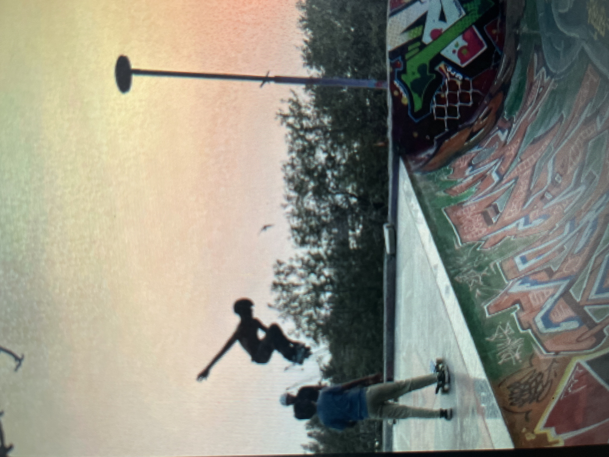 The image captures a dynamic scene at a skatepark in Sejahtera, Indonesia. In the foreground, a curved ramp is covered in vibrant graffiti. Beyond the ramp, a group of individuals are present. One person, silhouetted against a hazy, pastel sky, is captured mid-air performing a skateboard trick. Another person, wearing a blue shirt and light-colored pants, stands on the flat surface of the skatepark, facing away from the camera, with a skateboard at their feet. A third figure, also silhouetted, stands behind the person in the blue shirt. Trees form a dense backdrop to the scene, and a tall pole with a lamp at the top stands to the right. The overall lighting suggests it is either dawn or dusk, with a warm, diffused light illuminating the sky.