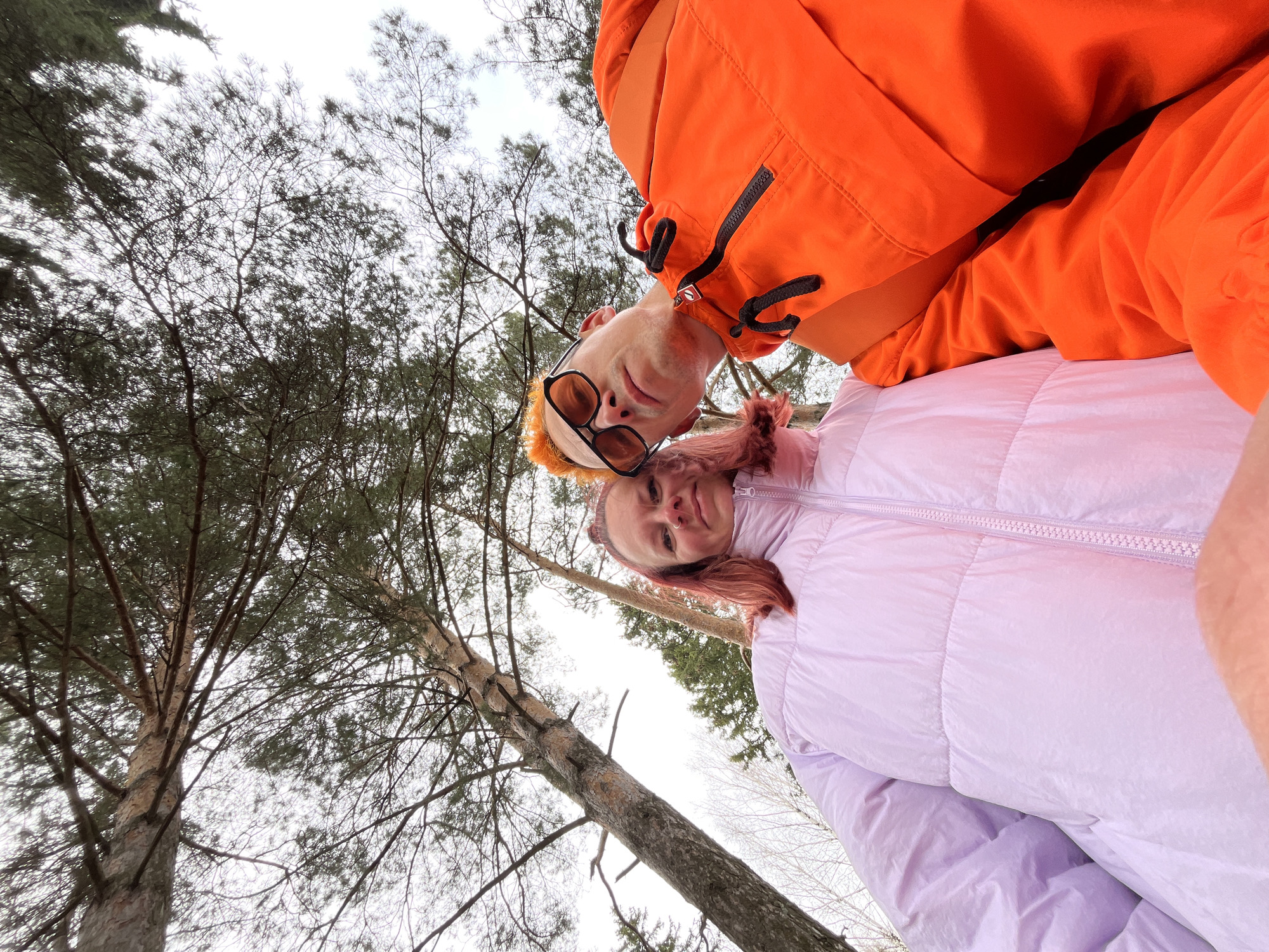 This unique, low-angle portrait captures two individuals against an overcast sky and a forest canopy in Chaikovsky, Russia. The image is rotated 90 degrees clockwise, suggesting it was taken from a very low perspective, possibly with the subjects lying down or holding the camera directly upwards.
In the foreground, on the left, a person with short, reddish-orange hair wears a bright orange hooded jacket with black zippers and drawstrings, and dark-rimmed sunglasses. They are looking upwards, towards the camera. To their right, a second person, with reddish/pinkish hair, wears a light lavender puffer jacket with a prominent zipper. This individual also looks towards the camera, displaying a slight smile or smirk.
The background is dominated by the tall, slender trunks and branches of numerous trees, primarily conifers with needle-like leaves, and some bare deciduous branches visible against the pale sky. The sky appears uniformly grey and overcast, indicating a cloudy day without direct sunlight. The subjects' warm jackets suggest cool weather, possibly late autumn, winter, or early spring. There is no visible text within the image. The overall impression is of a casual outdoor selfie taken during a leisurely moment in a wooded area.