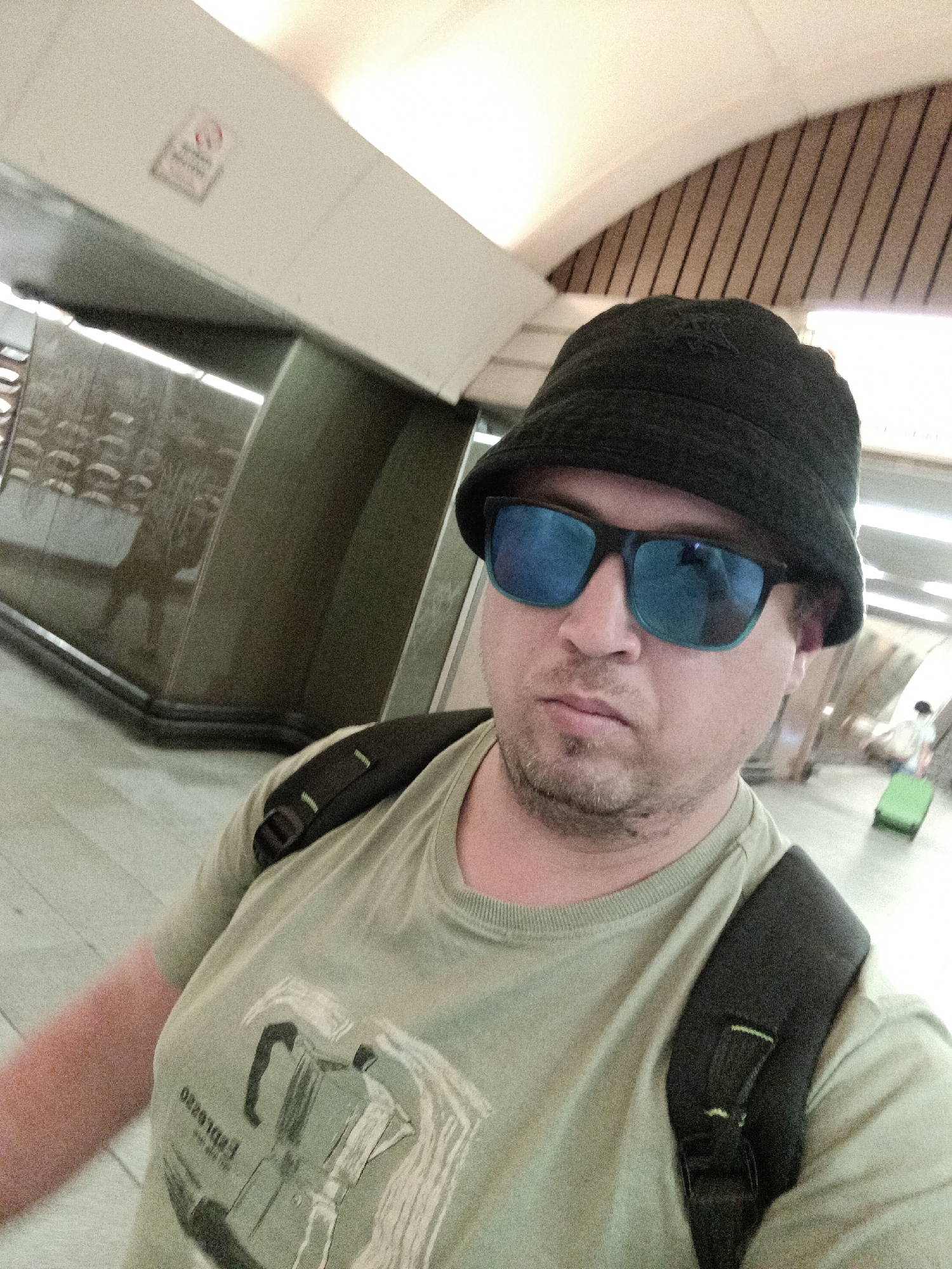 A man wearing a black bucket hat, blue mirrored sunglasses, and a light green t-shirt with a graphic print on it is taking a selfie. He is also wearing a black backpack. He is standing in what appears to be a public indoor space with a curved ceiling, tiled floor, and reflective walls. In the background, a person with a green suitcase can be seen walking away. A "no smoking" sign is visible on a light-colored surface on the left side of the frame. The overall lighting suggests it is daytime. The location is identified as Prague, Czechia.