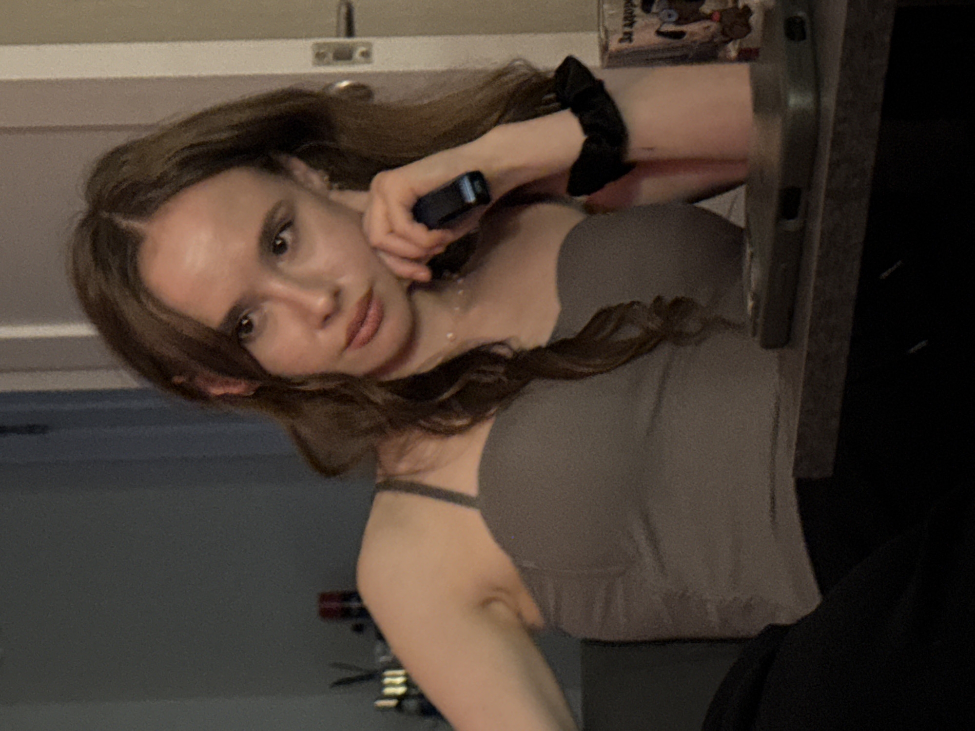 A young woman with long, brown hair is leaning towards a surface, possibly a table, with her head tilted. She is holding a dark-colored object, resembling a phone or small electronic device, up to her ear, as if engaged in a phone call. She is wearing a taupe-colored top with thin straps. A black scrunchie is visible on her wrist. In the background, there is a light-colored wall with what appears to be a light fixture or ventilation. Part of a dark object, possibly clothing, is visible on the right side of the frame, and a blurry object with some colorful elements is on a shelf behind her. The lighting suggests it might be indoors during the evening or night.