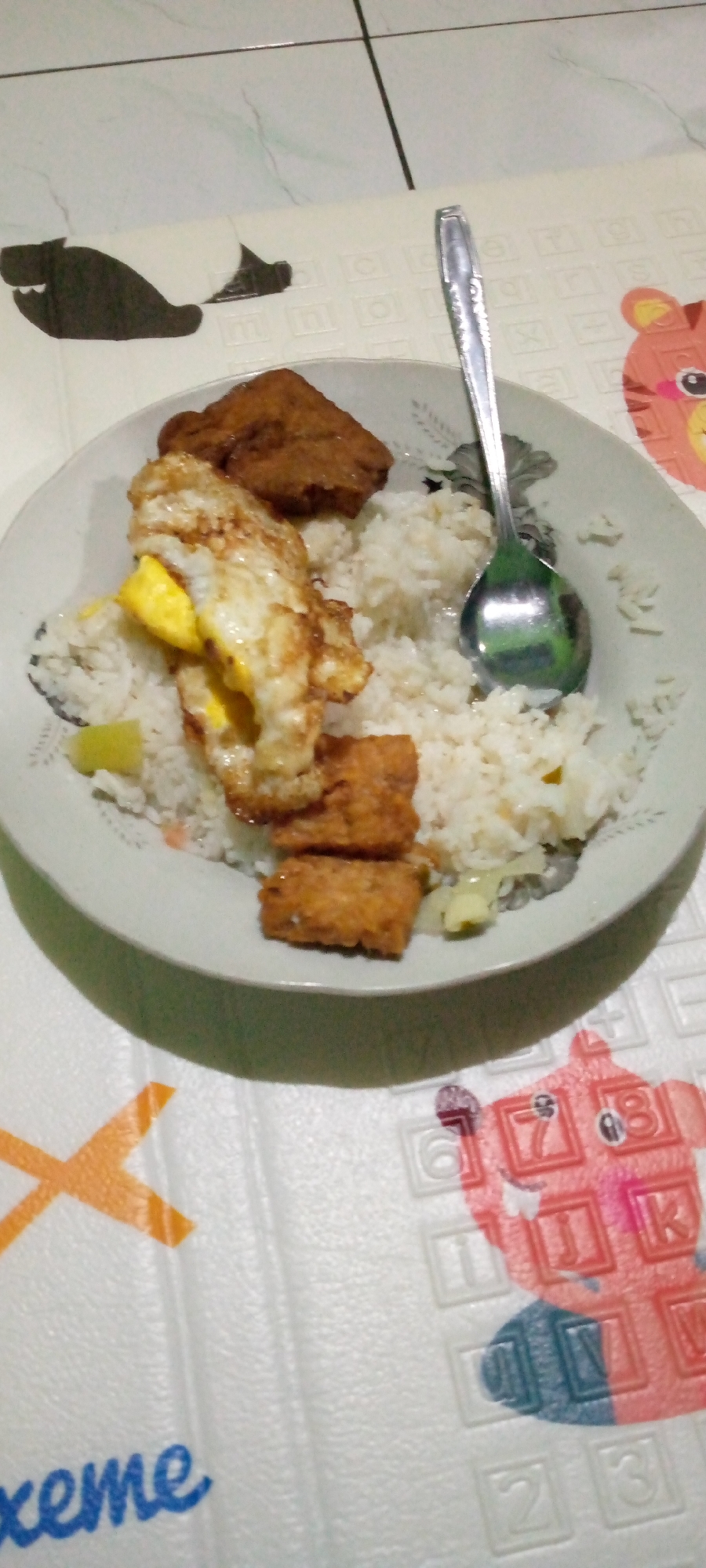 In the city of Sumberwono, Indonesia, a plate of food rests on a white surface with a cartoon animal print. The plate contains white rice, a fried egg with a bright yellow yolk, and several pieces of fried tempeh. A silver spoon is placed on the rice, its bowl reflecting the surrounding colors. The background features a tiled floor.