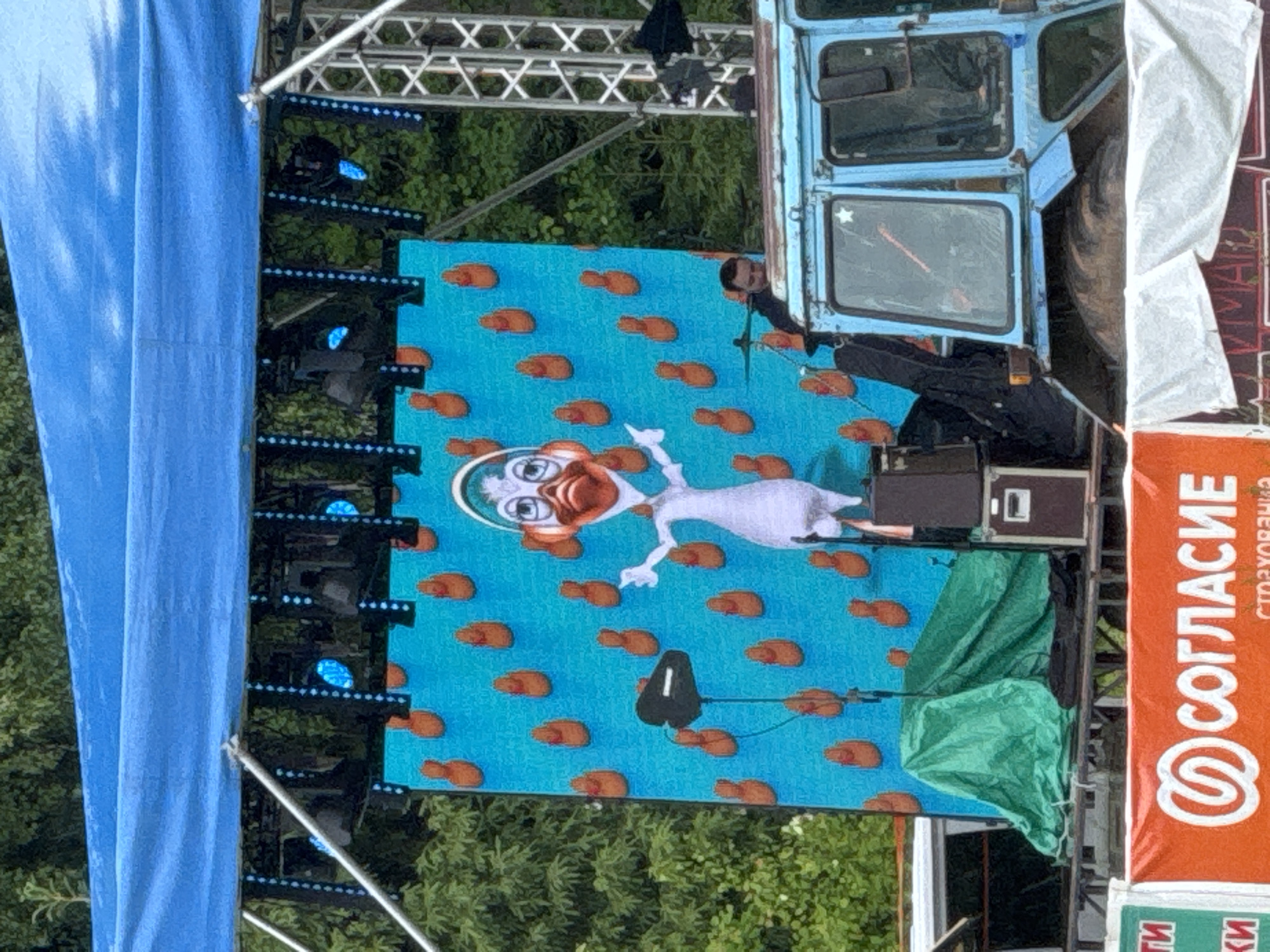 The image depicts an outdoor stage setup, likely for an event or concert, against a backdrop of trees. A large LED screen dominates the center, displaying an animated white duck or goose-like character with orange features and headphones, floating on a bright blue background speckled with numerous small, brown, shrimp-like or peanut-shaped objects.
To the left of the screen, a large blue tarp or curtain hangs vertically, partially obscuring metal truss structures equipped with stage lighting fixtures that emit blue light. To the right of the screen, the front cabin of an old, light blue vehicle, possibly a truck or bus, is positioned. A person with dark hair is partially visible looking out from the vehicle's window, and another person's arm and shoulder are seen near the bottom right of the screen, next to sound equipment. Below the vehicle's cabin, sound equipment, including a speaker or amplifier, is covered with a green tarp.
On the far right, a vertical orange banner is prominently displayed, featuring the text "СОГЛАСИЕ" (SOGLASIE) and "СТРАХОВАНИЯ" (STRAKHOVANIYA), which translates to "CONSENT" or "AGREEMENT" and "INSURANCE" respectively, indicating a sponsor or organization. A smaller green banner with partial text "ТИ" and "КИ" is visible below the orange one.
The scene appears to be taking place during the daytime, given the natural light on the foliage, although the stage lights are active. The overall impression is that of a public outdoor event, possibly family-oriented given the animated character on the screen. The Russian text on the banner confirms the location context of Bol'shaya Kivara, Russia.