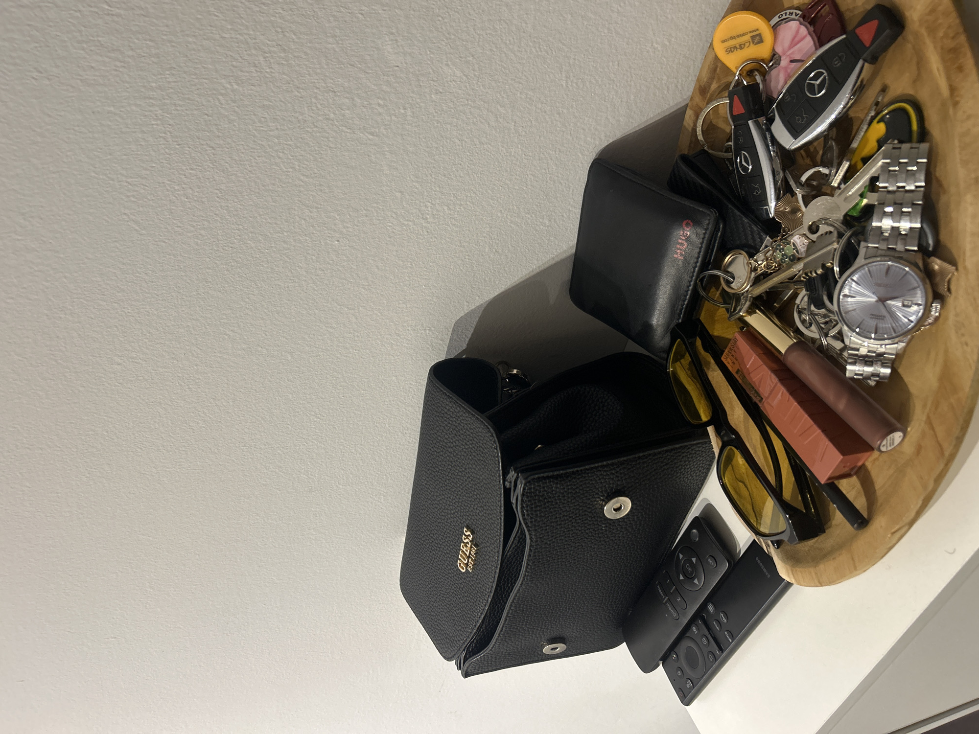 The scene, situated in the city of Varna, Bulgaria, depicts a collection of personal effects arranged on a white flat surface, likely a table or shelf, against a light-colored textured wall. On the left, a black Guess handbag, featuring a gold "GUESS EST.1981" logo and magnetic snap closures, stands partially open. Two black television remote controls, one branded Samsung, rest on the surface adjacent to the handbag. To the right, a shallow, light-brown wooden bowl contains various items. Within the bowl, a black Hugo Boss wallet with a red "HUGO" logo is visible beneath a dense cluster of keys. This cluster includes multiple house keys, two Mercedes-Benz car key fobs, a yellow circular "LONGS" tag, and several decorative keychains. A silver-toned metallic wristwatch with a light-colored dial is also present in the bowl. Alongside these items are a pair of sunglasses with black frames and yellow-tinted lenses, and two tubes of lip product, one orange-brown and one darker brown. Shadows are cast on the wall behind the objects, indicating an indoor setting with a light source from the upper left.