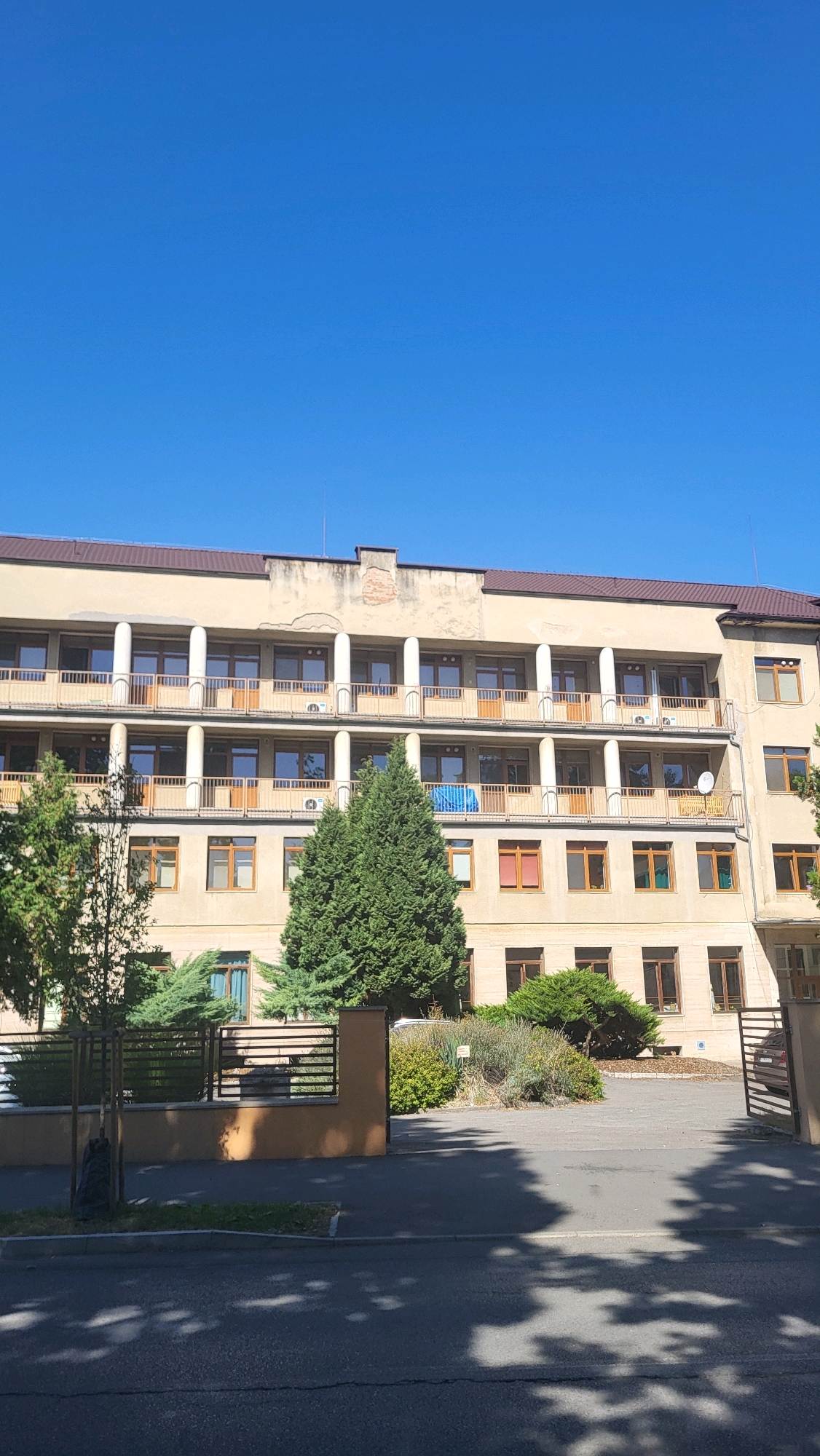 This is a photograph of a light-colored, multi-story building with multiple balconies and windows, set against a clear blue sky. The building appears to be a hospital or similar institution, evidenced by the architectural style and the presence of what seem to be air conditioning units visible on some of the balconies. Trees and shrubbery surround the building, and a fence is visible in the foreground, partially obscuring the view of the building's lower levels. Shadows from trees are cast across the pavement in front of the building, suggesting a sunny day. The building is likely located in Prešov, Slovakia, based on the request for location-specific information. There are no people or animals visible in the frame.