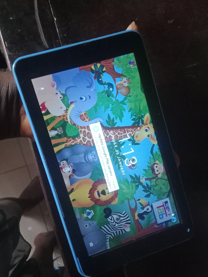 A tablet with a blue frame displays a children's cartoon-themed lock screen. The wallpaper features an elephant, giraffe, lion, zebra, monkey, and other animals in a jungle setting. A notification window prompts the user to update Google Play Services, indicating that certain apps won't function correctly without the update. The screen shows the time as 1:13 and the date as Sunday, January 25. A "Legacy Tab" icon is visible in the corner.