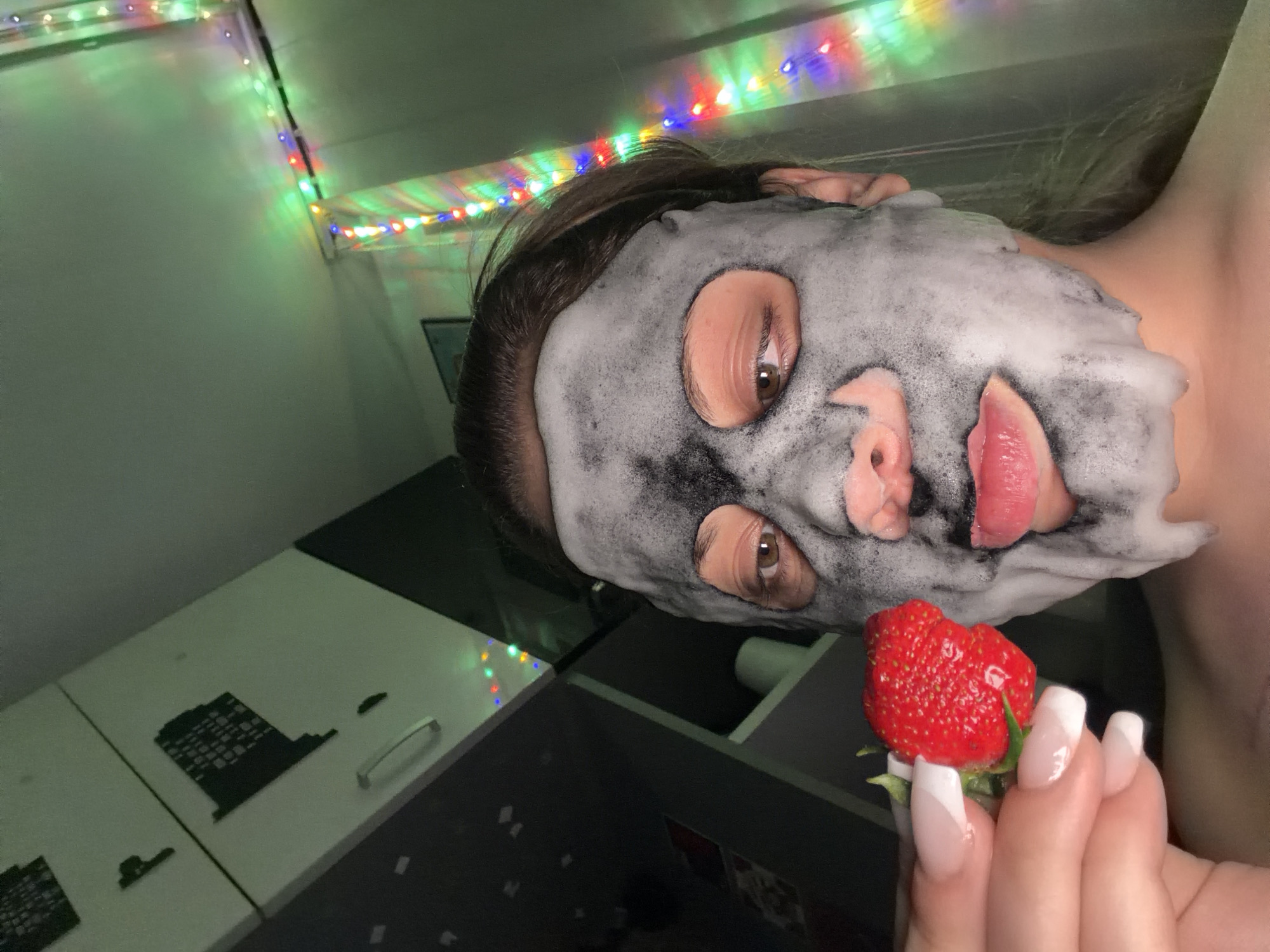 A close-up, angled shot shows a person with a foamy gray facial mask, holding a ripe red strawberry with green leaves near their face. The person's right hand, displaying long, white-tipped fingernails, grasps the strawberry. Colorful fairy lights are visible along the upper edge of the image. The background includes white and black cabinet doors. The location is uncertain based on the image; it is unknown if it is in Varna, Bulgaria.