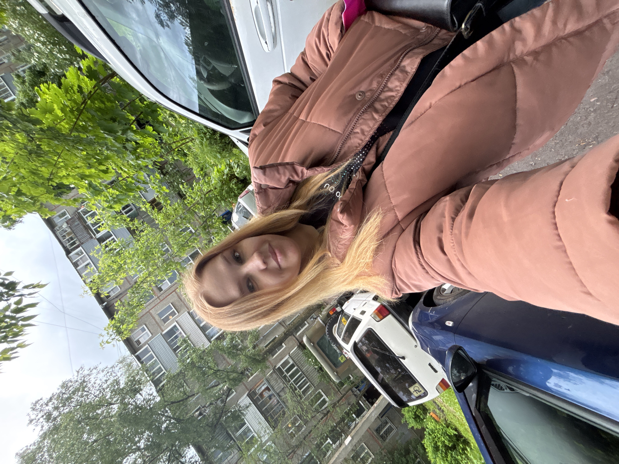 The image shows a blonde woman posing in front of a building in Заволжье, Russia. She is wearing a brown puffer jacket. Parked cars are visible on the street. The trees are green, indicating it is likely spring or summer. The building has many windows.