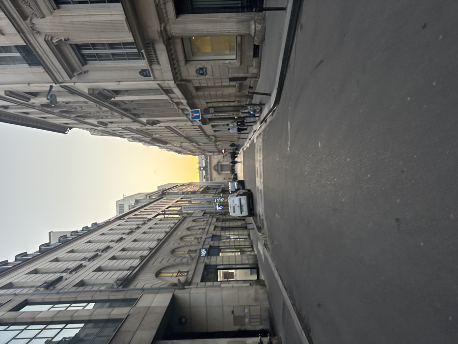 The image displays a perspective view down Rue Notre Dame des Victoires in the 2nd arrondissement of Paris, France, looking westward at what appears to be late afternoon or early evening. The street, paved with asphalt, recedes into the distance, flanked by multi-story Parisian buildings on both sides. The sky above transitions from a pale blue at the zenith to a vibrant orange and yellow gradient near the horizon, indicating a low sun angle.
On the left, buildings feature classical stone facades with modern glass elements on lower floors, housing retail establishments, one with a visible "Bodys" sign. A white delivery van is parked on the left side of the street, partially on the sidewalk. On the right, the buildings also exhibit traditional Haussmannian architecture with stone facades, numerous windows, and decorative elements. Street signs indicating "RUE DE LA BANQUE" and "RUE NOTRE DAME DES VICTOIRES" are affixed to the rightmost building. A blue "no through road" or "dead end" style sign is also visible on the right sidewalk, which features multiple black bollards and a public waste bin.
In the far distance, a prominent, ornate archway marks the entrance to a building or covered passage, likely leading towards the Passage des Panoramas complex. Several pedestrians are visible further down the street, mostly silhouetted against the brighter background, walking along the sidewalks and crossing the road. The street surface shows some reflective patches, possibly from recent moisture.
