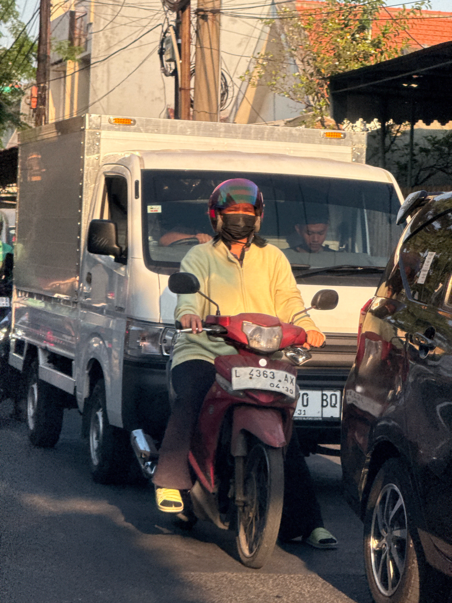 In Surabaya, Indonesia, on a city street, a person wearing a yellow jacket, a black mask, and a iridescent helmet rides a red motorcycle. The motorcycle has a license plate that reads "L 4363 AX 04-30" and a portion of another plate "0 BQ". The rider is positioned between a silver truck and a dark-colored car. The truck is a box truck with a silver cargo area and a white cab. The car is a dark sedan. Sunlight casts shadows on the road, indicating it is likely late afternoon. Buildings and power lines are visible in the background.