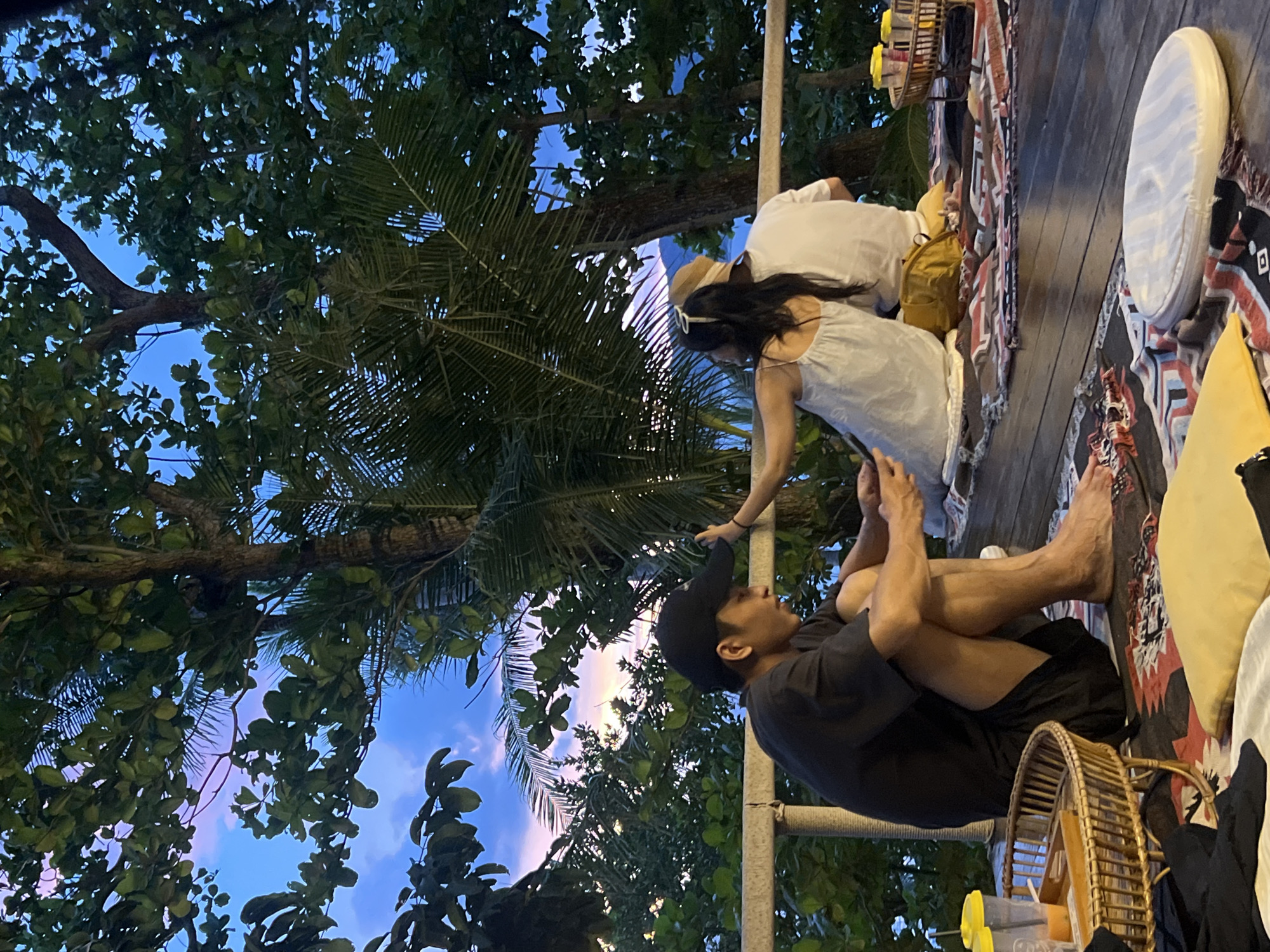The image shows a group of people outdoors, likely in a tropical setting, as indicated by the lush green trees and palm fronds in the background against a twilight sky. Two individuals are seated on a wooden deck, adorned with colorful textiles and cushions, suggesting a relaxed gathering or seating area. One person, wearing a black t-shirt and a baseball cap, is sitting cross-legged, looking upwards. Another person, dressed in white, is partially visible, leaning forward. A third person, also in white and wearing a straw hat, is seen from behind. The lighting suggests it is late evening or early morning, with soft light filtering through the trees. The overall atmosphere appears serene and casual, possibly a moment of rest or conversation during an outdoor event.
