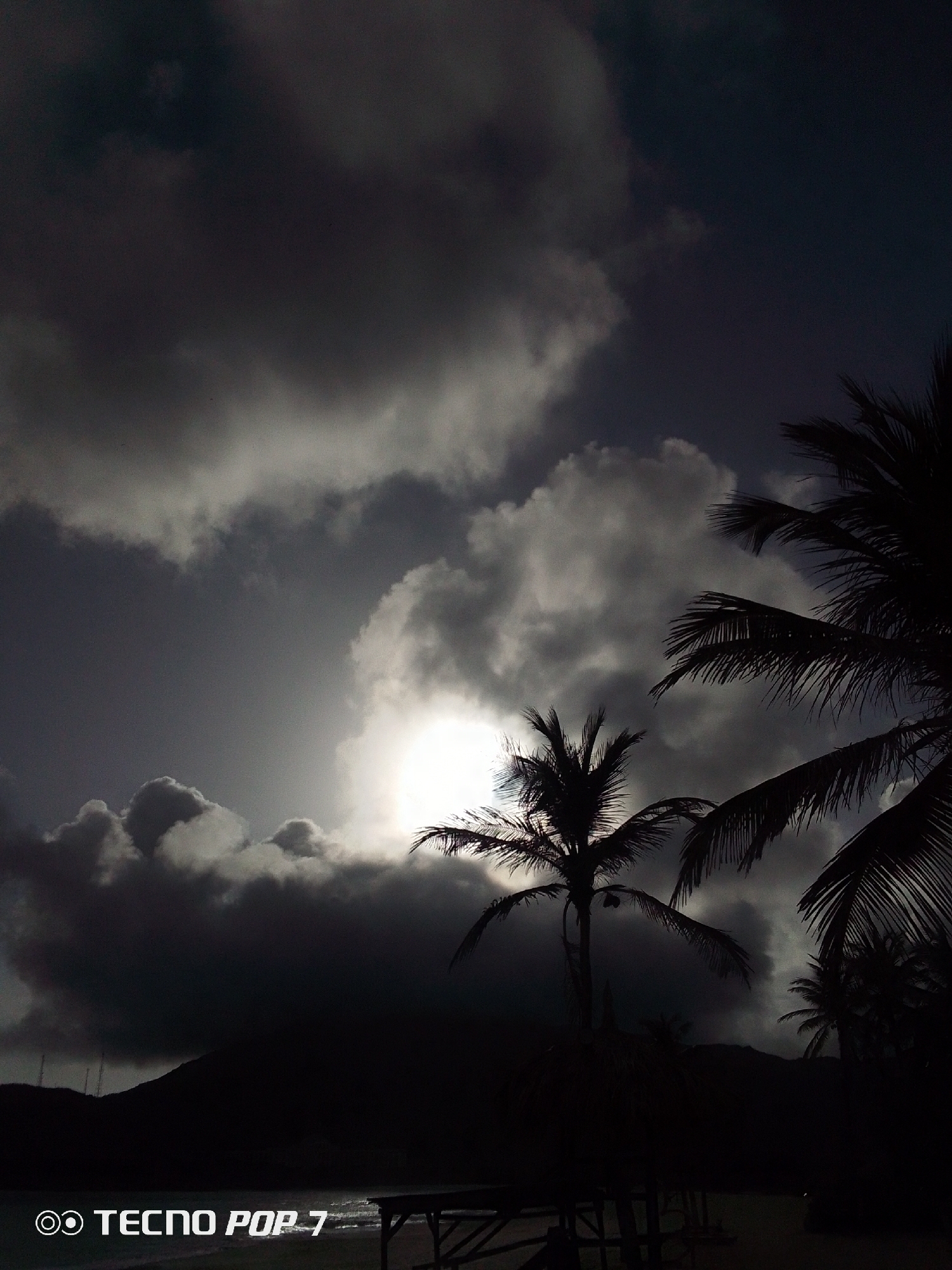 The image depicts a beach scene in Pedro González, Venezuela. The foreground contains a partially obscured wooden structure, possibly a picnic table. Two palm trees are silhouetted against a cloudy sky. The sun is visible behind a dense cloud cover. In the background is a dark land mass near the ocean. The image appears to be taken at dusk or dawn. The logo "TECNO POP 7" is visible at the bottom of the image.