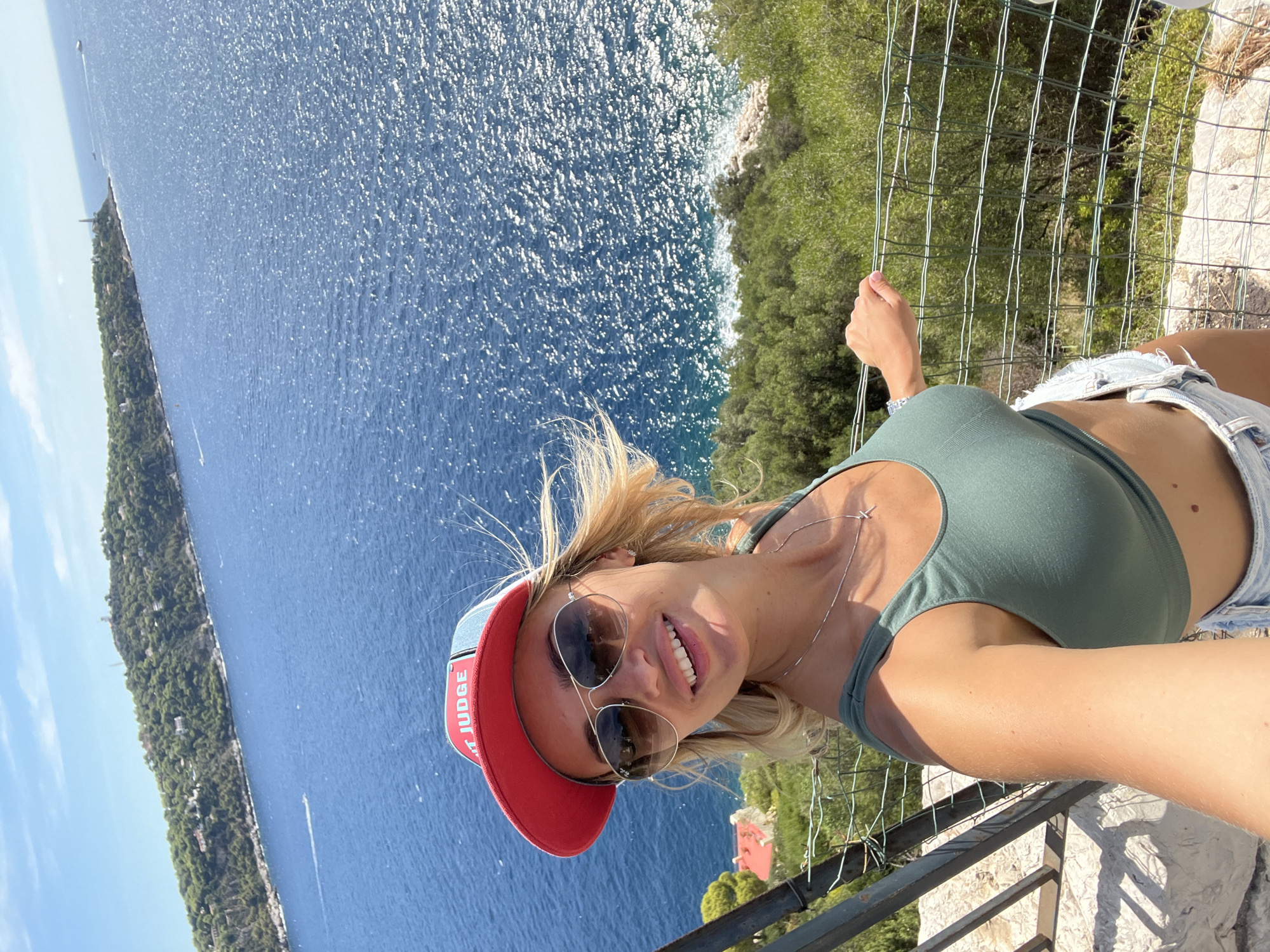 A woman with blonde hair and sunglasses poses for a selfie on a balcony overlooking the Mediterranean Sea. She is wearing a green crop top, denim shorts, and a red and blue baseball cap. The sea is a deep blue, with sunlight glinting off the surface. In the distance, a green, tree-covered island juts out into the water. The woman appears to be in Nice, France, given the coastal scenery and architecture visible in the background.