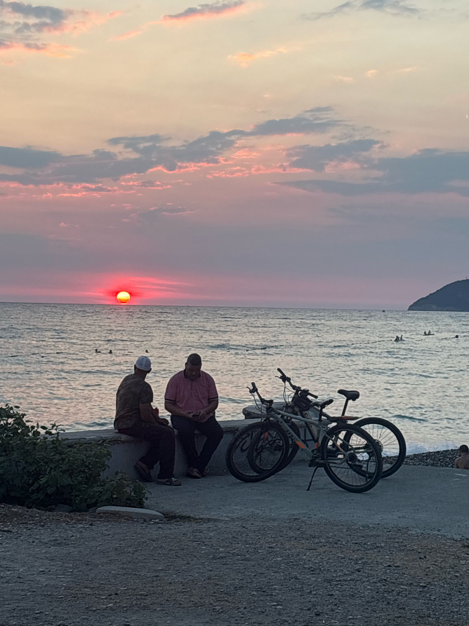 Two men are sitting on a concrete ledge facing the sea in Bar, Montenegro, during sunset. The sun, a bright red orb, is partially obscured by the horizon and reflected in the calm sea. The sky is filled with soft pink and orange clouds, transitioning to a pale yellow near the horizon. To the right, two bicycles are parked on the concrete promenade. The sea is gently rippling, and several people are visible swimming or wading in the water in the distance. A dark, leafy bush grows to the left of the men. The ground in the foreground is a mixture of gravel and concrete. The overall scene depicts a peaceful evening by the Adriatic Sea.
