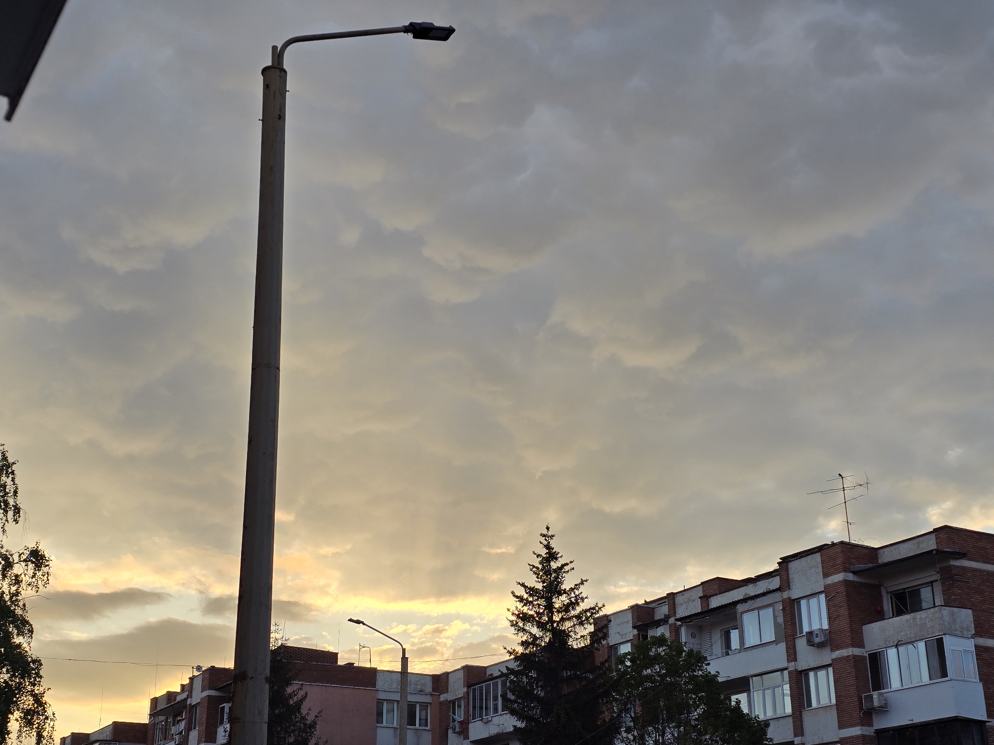 This image captures an urban or suburban scene at either sunrise or sunset, dominated by a dynamic sky filled with clouds. The upper two-thirds of the frame are covered by a thick layer of grey and blue-grey altocumulus and stratocumulus clouds. Towards the horizon, particularly on the left, the clouds become less dense, allowing golden and yellow light from the setting or rising sun to filter through, illuminating the undersides of the clouds with warm tones.
A prominent, tall, grey, cylindrical metal streetlamp stands on the left side of the frame, extending from the bottom to near the top. It is equipped with a modern, dark, rectangular LED light fixture. Another, smaller streetlamp can be seen further back, just above the buildings on the left, also silhouetted against the sky.
In the mid-ground and background, a row of multi-story residential buildings is visible. These structures feature a mix of materials, including red-brick sections and lighter-colored (white or grey) facades. Numerous windows and balconies, some of which are enclosed, are apparent on the building exteriors. Several air conditioning units are mounted on the walls. On the roof of the building to the right, a television antenna is clearly visible.
Trees are also part of the landscape; a dark, conifer tree, likely a pine or fir, stands out against the lighter sky in the mid-ground, and parts of leafy trees are seen on the far left edge of the image. The overall mood is calm, evoked by the soft, diffused light and the cloud-filled sky. There are no people or discernible text in the photograph.