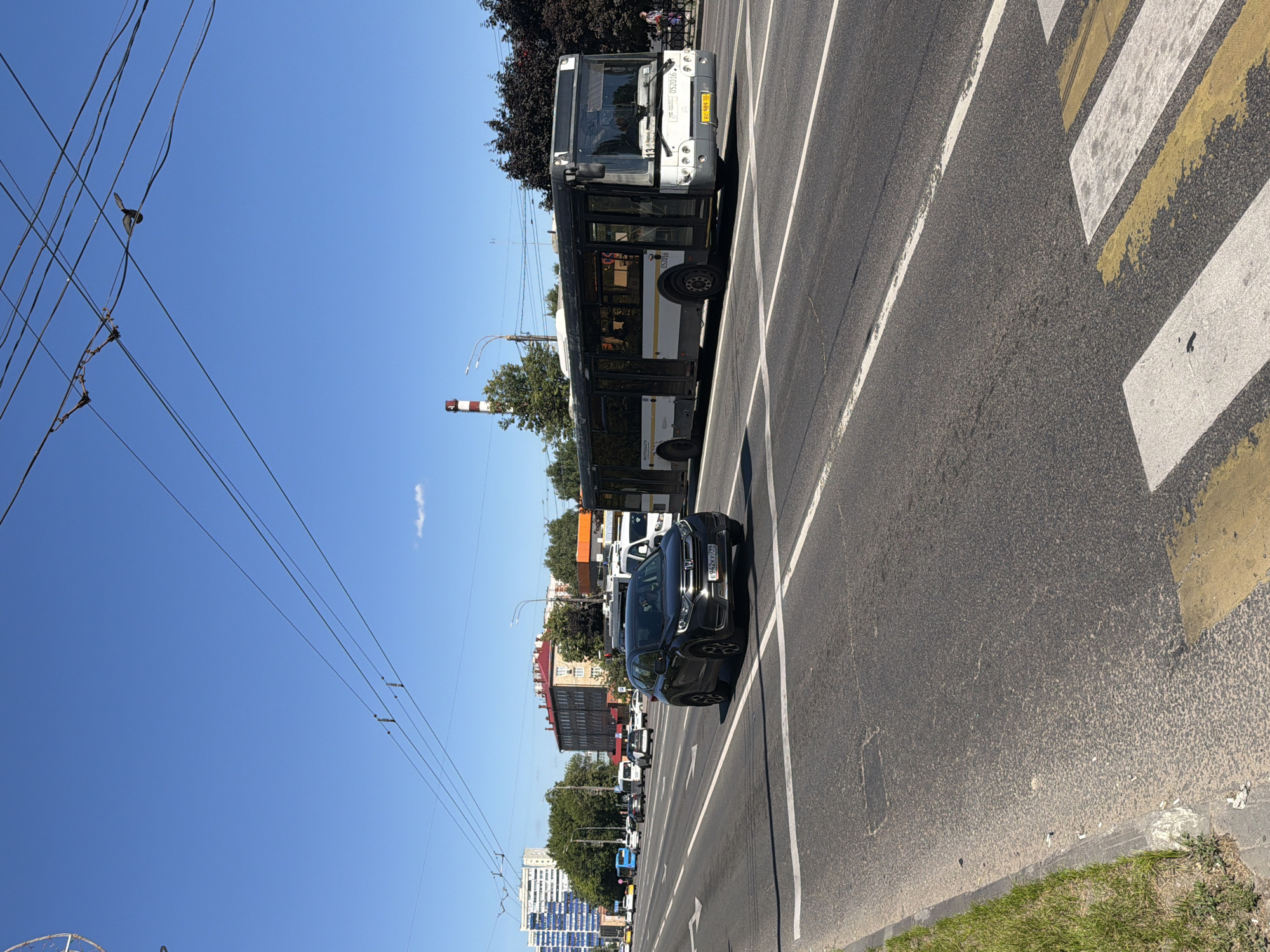 The image depicts a wide street in Podolsk, Russia, under a clear blue sky. A dark grey SUV is stopped at a pedestrian crossing in the foreground, facing right. Behind it, a long, articulated bus, predominantly white with black windows and trim, is moving on the right lane of the road. The bus has the route number "13" visible on its front. The street has multiple lanes of traffic, marked with white lines. Various buildings, including multi-story residential blocks and what appears to be a factory chimney with red and white stripes, are visible in the background. Trees line the sides of the road, and overhead power lines stretch across the frame. Several other vehicles, including buses and cars, are present further down the street. The scene appears to be a typical urban environment on a sunny day.