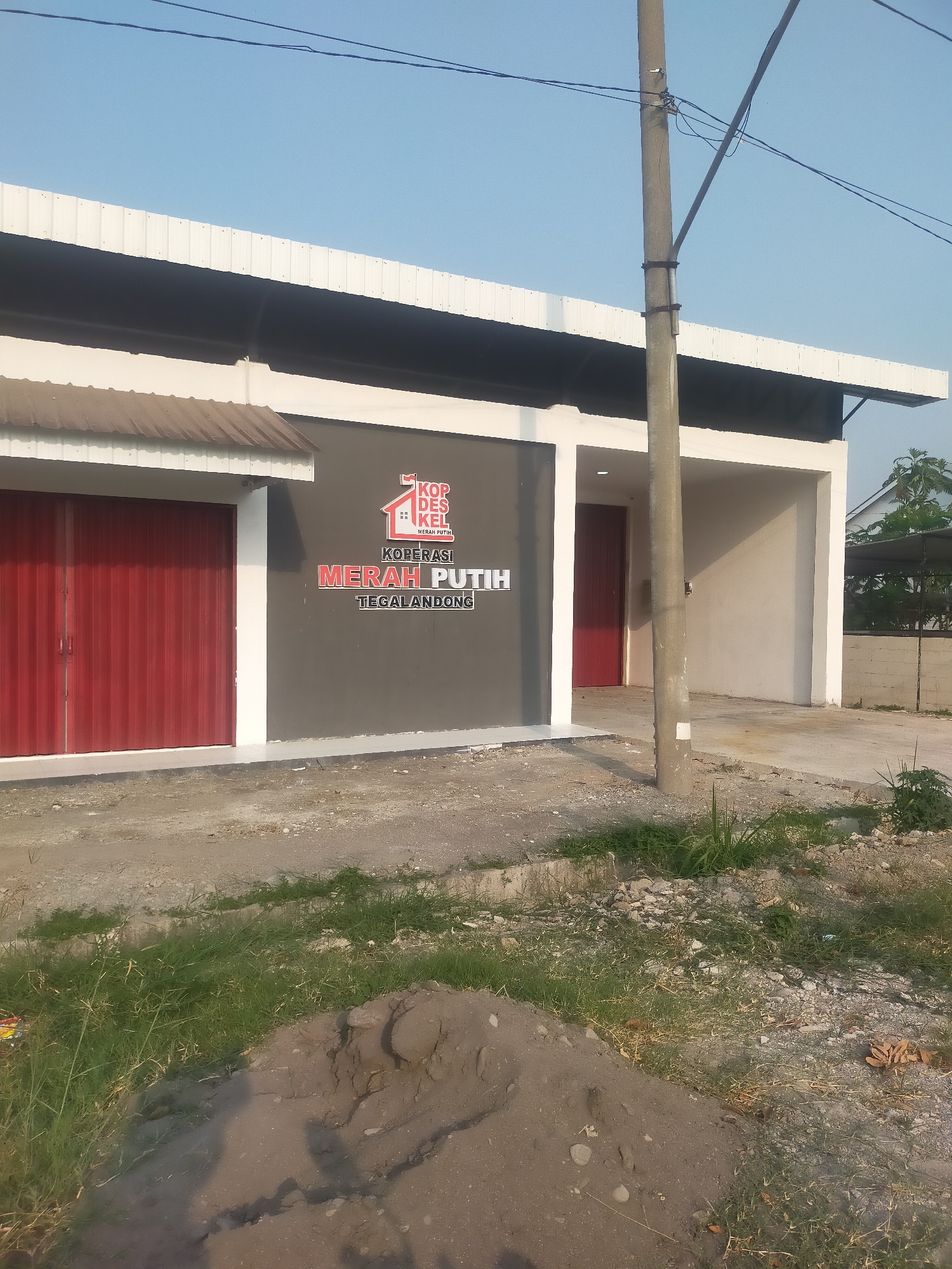 The image shows the exterior of a building with a dark gray facade and red roll-up doors. A logo with text that appears to be in Indonesian is displayed on the facade. The building is situated in a semi-urban or rural environment, with a dirt road and grass in the foreground. A utility pole with wires is also visible. The sky is clear and blue. The building appears to be a commercial establishment, possibly a cooperative, given the text on the logo. The overall impression is of a new or recently constructed building in an undeveloped area.