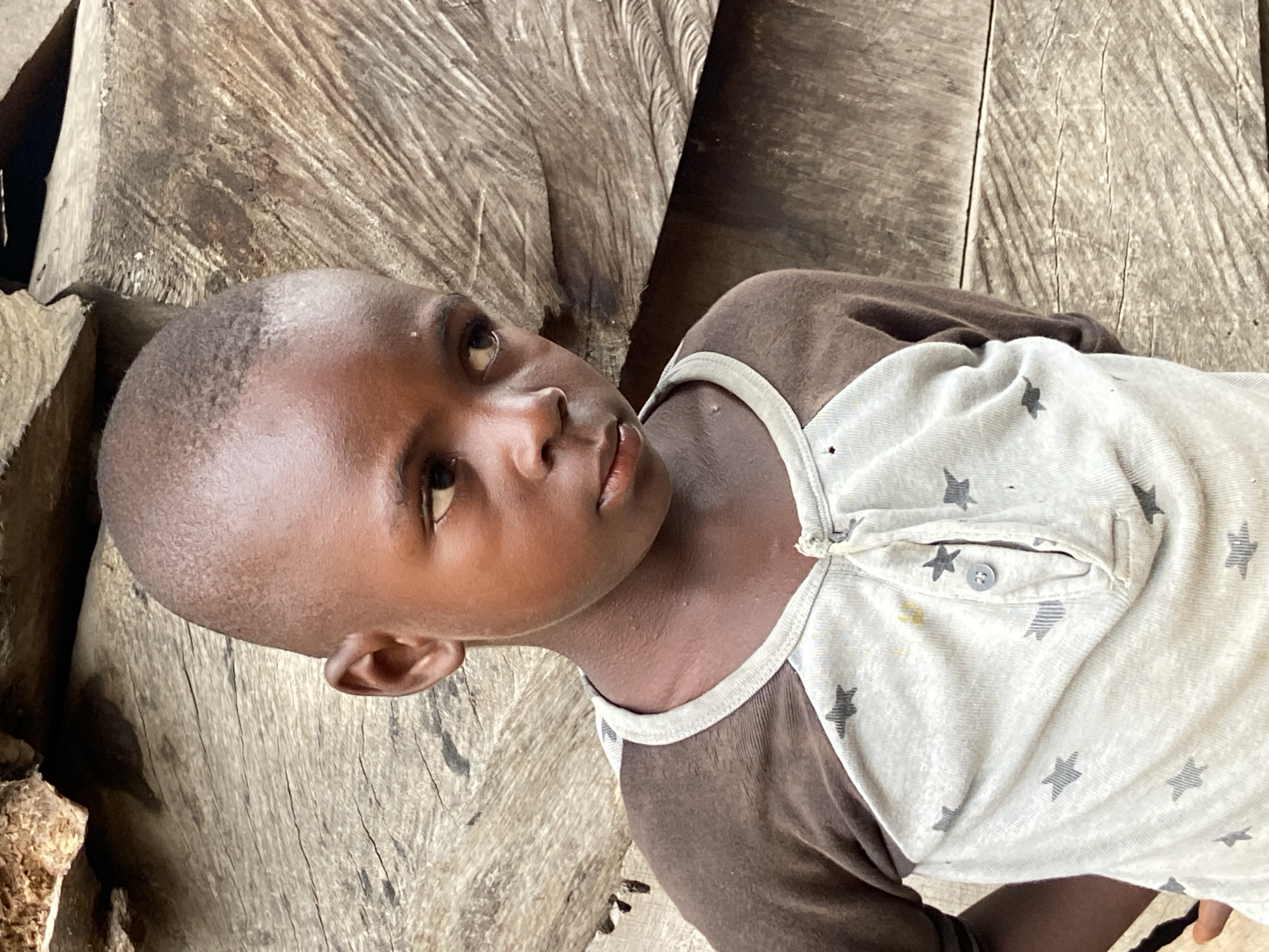 A young, dark-skinned boy is lying on a rough, weathered wooden surface. He is wearing a brown and white patterned shirt with stars. His expression is thoughtful, and he is looking off to the side. The scene appears to be outdoors, with the wooden surface possibly a part of a rough structure. There is no visual information to confirm the location is in Mararaba, Nigeria.