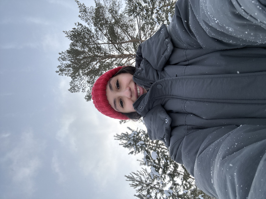 A close-up, low-angle selfie captures a young female subject outdoors in Sysert, Russia. The subject wears a red knitted beanie and a dark grey, puffy winter coat, which shows scattered snowflakes. She is smiling, with teeth visible. The background consists of an overcast, light grey sky and a scattering of snow-covered evergreen trees, primarily pine or fir. Smaller trees with accumulated snow on their branches are visible on the left, while a taller, more sparsely snow-dusted pine tree stands on the right. Visible flakes in the air indicate ongoing snowfall.