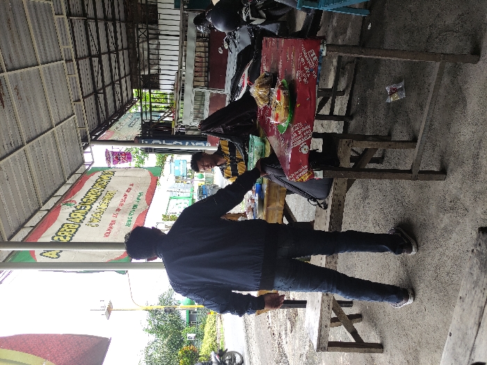 This image depicts a casual, outdoor dining scene in Kepanjen, Indonesia, likely at a street-side eatery or *warung*. The setting is characterized by a simple structure with a corrugated metal roof, providing shade over a concrete ground.
Two men are visible in the frame. In the foreground, a man stands with his back mostly to the viewer, dressed in a dark blue jacket, blue jeans, and sneakers. He appears to be either approaching or interacting with another man seated at a wooden table in the mid-ground. The seated man is wearing a yellow and black striped shirt, and his face is partially visible as he looks towards the foreground.
On the wooden table, which is covered with a red material featuring small, repeated white text, there is a plate holding food, possibly noodles, and a small plastic bag containing snacks. Another wooden chair is pulled up to the table. In the background, a motorcycle is partially visible behind the seated person.
A large, light-colored banner with green borders is prominently displayed, hanging from the roof structure. The text on the banner is partially mirrored and obscured, but discernible words include "BEBEK" (Indonesian for duck) and possibly "SUPER GUR...", suggesting it advertises a food stall specializing in duck dishes. Beyond the immediate structure, trees and a glimpse of what appears to be a road indicate an open-air environment.
The time of day appears to be daytime, with bright but not harsh lighting, suggesting clear weather. The overall atmosphere is relaxed and commonplace for a local eatery in the region.