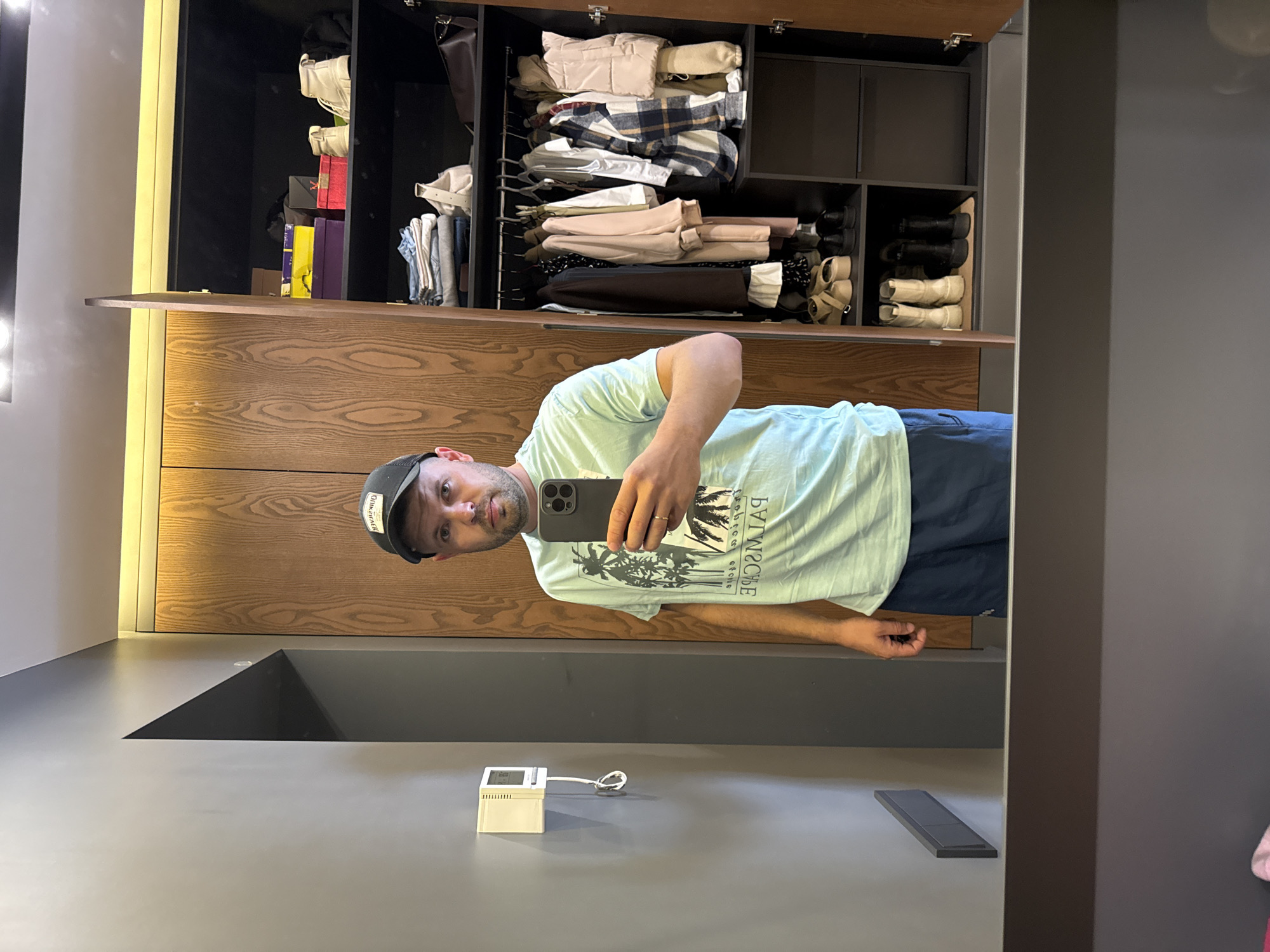 The image shows a man in a light blue t-shirt and navy blue shorts taking a selfie in a mirror. He is wearing a black baseball cap. Behind him is an open closet with various clothes and shoes neatly organized on shelves and hanging on a rack. The closet doors are made of wood. To the left of the closet is a gray wall with a white thermostat. The scene appears to be indoors, possibly in a bedroom or dressing area. The presence of clothing and personal items suggests a domestic setting. The image does not provide any specific visual cues to identify the location as Novolisiha, Russia.