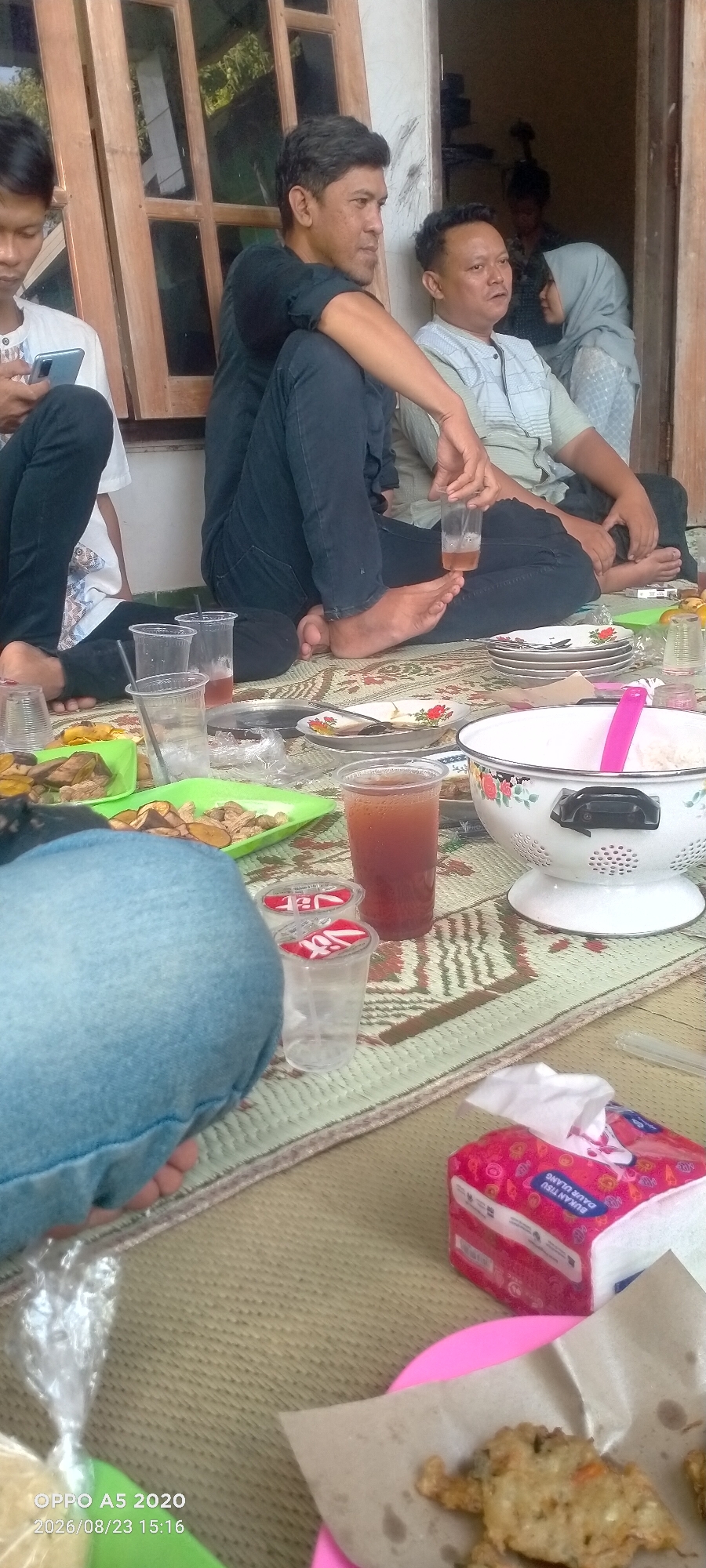 In Sendang, Indonesia, a group of individuals are seated on the floor, likely on a mat, engaged in a casual gathering. The setting appears to be outdoors or in a semi-open space, with a wooden door frame and partial view of a building in the background.
In the foreground, several plastic cups containing beverages, one with ice and another with a darker liquid, are placed on the mat. Plates with food, possibly snacks or a meal, are also visible. One plate holds what appears to be fried food. A white bowl with a pink utensil is also present, possibly for serving food. A packet of tissues with a red and white design is in the lower right corner. The flooring is a woven mat with a patterned design.
Several people are the focus of the image. On the left, a person wearing a white shirt is looking down at a mobile phone. Next to them, a man in a black shirt is seated with his legs crossed and is looking to the right. Another man, wearing a light-colored patterned shirt, is seated next to him, also looking to the right. In the background, two more individuals are visible, one wearing a light blue headscarf, suggesting a culturally conservative environment. The overall atmosphere suggests a relaxed and informal social gathering.