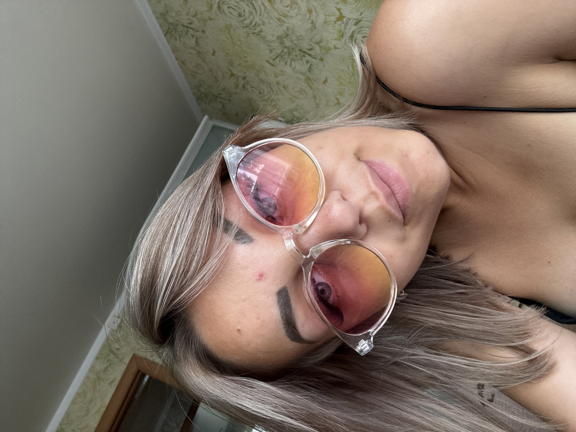 A young woman with blonde hair and sunglasses is pictured in a close-up, head and shoulders shot. She is looking towards the camera, with a slight smile on her face. The sunglasses have clear frames and lenses that transition from pink to orange. There is a small red blemish on her forehead. The background appears to be a wall with patterned wallpaper and a portion of a ceiling. The lighting suggests an indoor setting, possibly a residential room. No specific location in Krasnogorskoye, Russia, can be identified from the visual information provided.