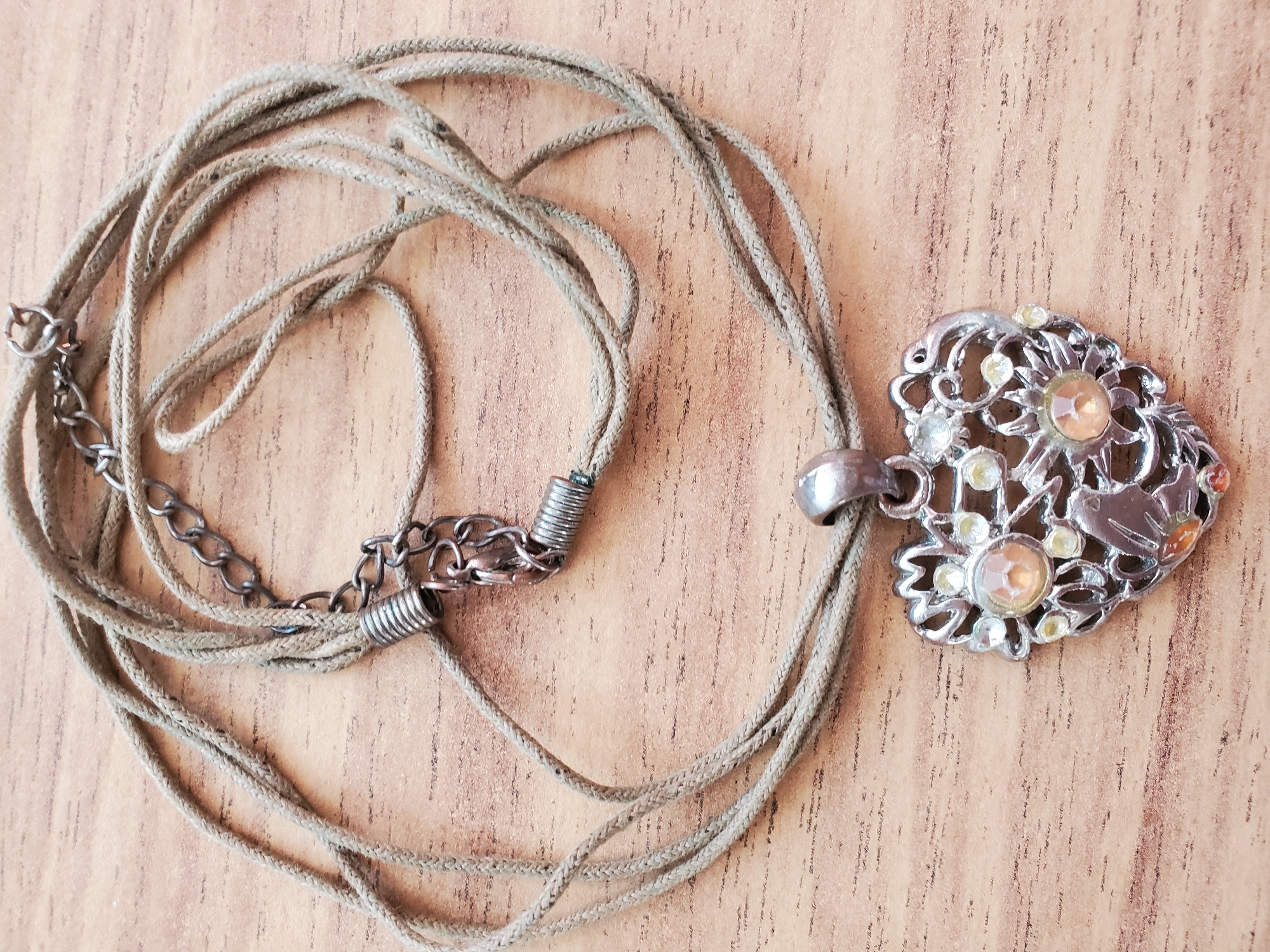 A close-up, overhead shot displays a silver heart-shaped pendant adorned with small, clear and amber-colored stones, attached to a thin, braided tan cord necklace. The necklace is looped loosely on a light brown wooden surface, exhibiting a subtle grain pattern. The pendant itself features intricate scrollwork and a central, larger amber-colored stone surrounded by smaller clear stones. A short silver chain with a clasp is visible, connecting to the cord. There are no people present in the image, and no contextual clues suggest the location is Tiraspol, Moldova.