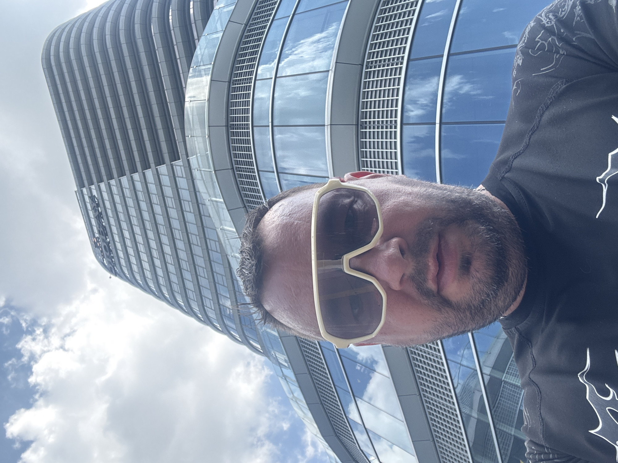A selfie is taken from a low angle, with the top of the photographer's head and face prominently featured in the foreground. They are wearing large, light-colored sunglasses and a black t-shirt. Behind them, a modern, curved glass building dominates the background, its reflective surface mirroring the cloudy sky above. The building's architecture features horizontal banding on its upper floors. The setting appears to be an urban environment, possibly the Suzhou Industrial Park in China, given the contemporary architectural style of the building and the overall urban ambiance. The sky is partly cloudy, with white clouds visible against a bright blue backdrop.