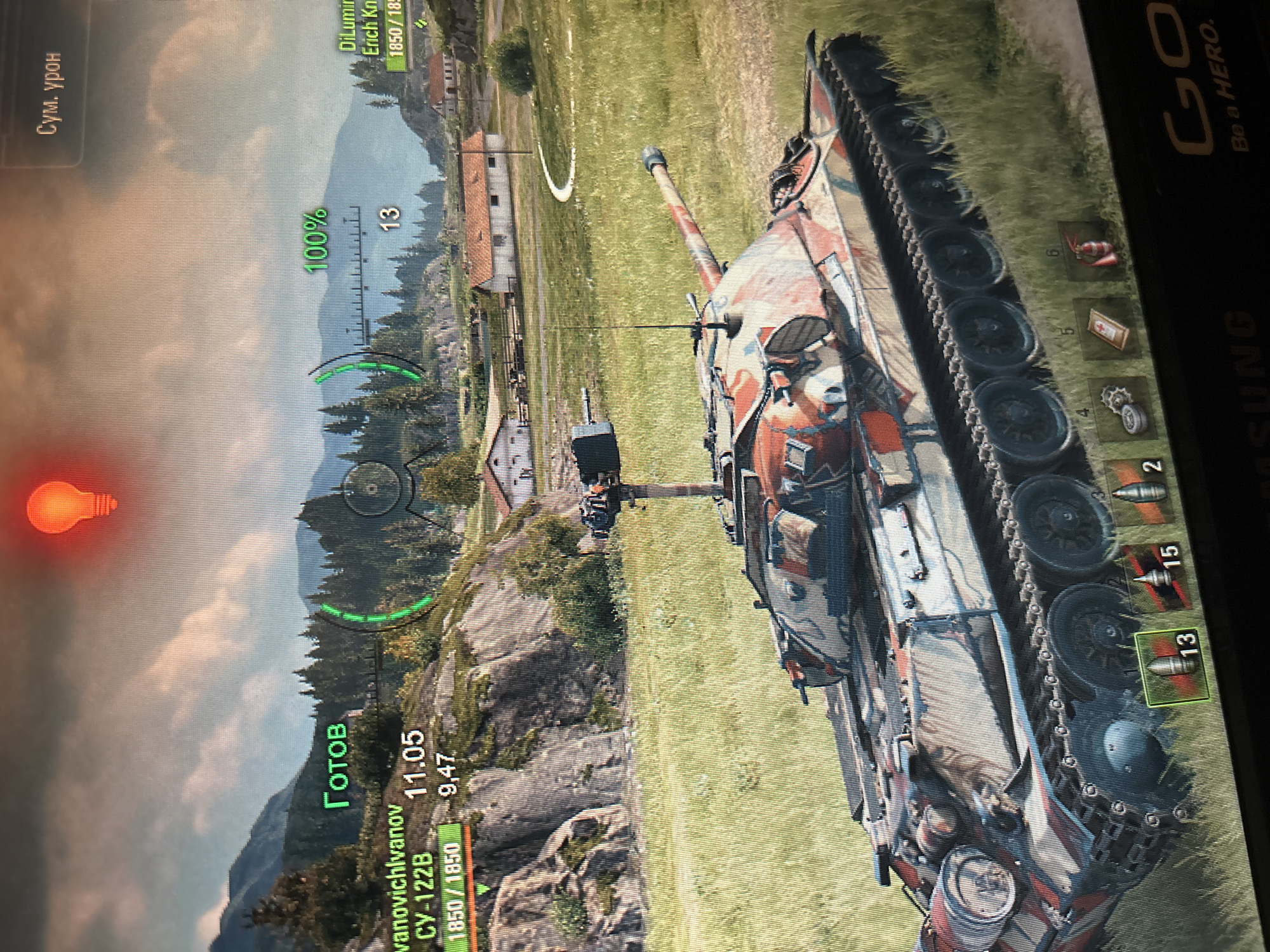 This is a screenshot from a video game, likely World of Tanks, showing a third-person perspective of a tank in a rural setting. The tank, a Soviet SU-122B, is camouflaged with red, white, and blue patterns and is positioned in a grassy field with rolling hills and trees in the background. There are several buildings that appear to be farmhouses or rural dwellings scattered across the landscape. The sky is overcast with some clouds. The user interface elements are visible, including the tank's health (1850/1850),ammunition count (13), and a targeting reticle. Other icons indicate consumables like a medkit, fire extinguisher, and repair kit. The game's map shows the player's location and other players' general positions with numerical indicators. The overall scene is one of a virtual battlefield during a daytime match. There are no people visible in the image.