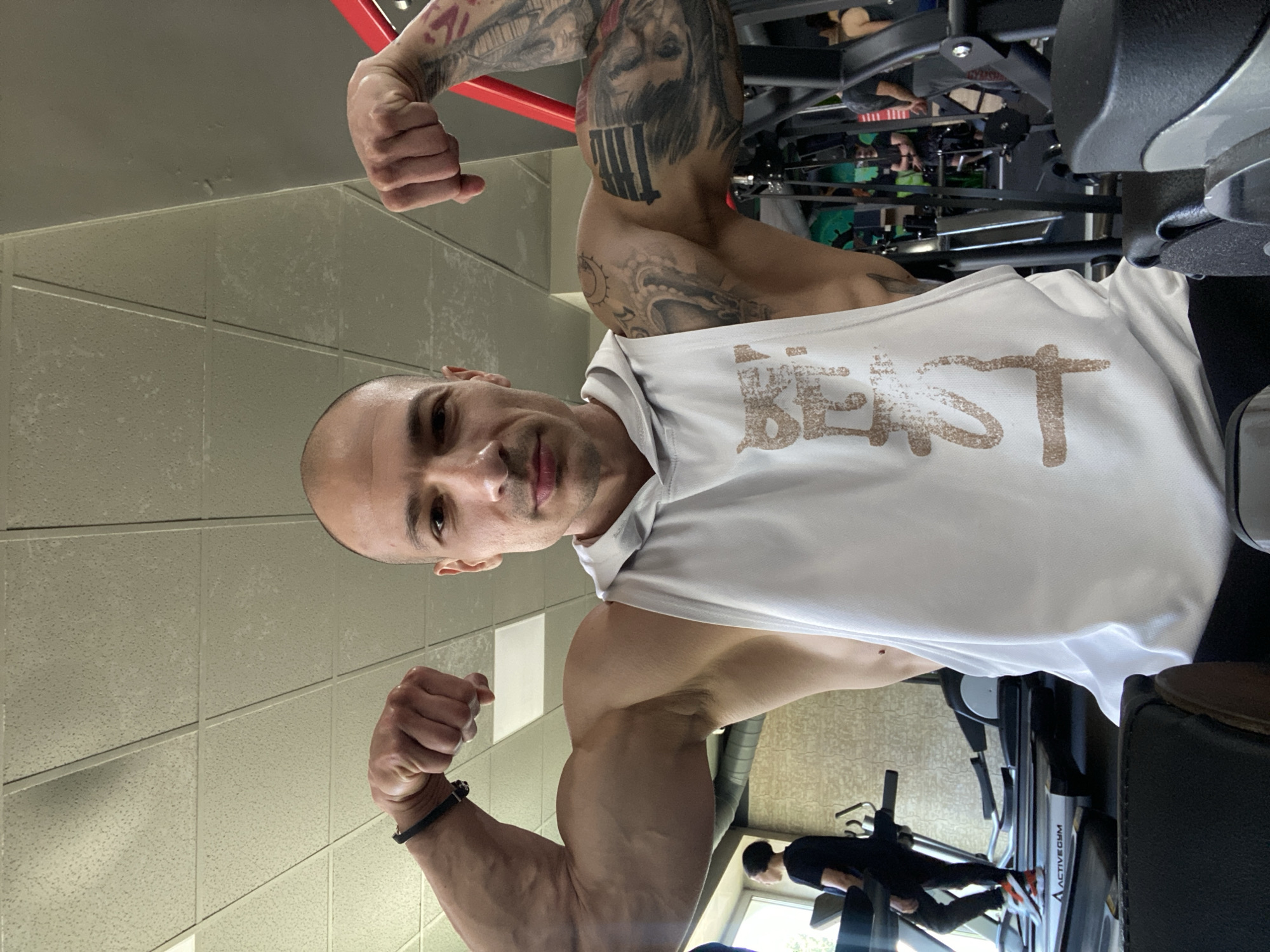 A man is posing and flexing his biceps in what appears to be a gym. The man has a shaved head and is wearing a white sleeveless shirt with the word "BEAST" printed on it. He has tattoos on his arms. In the background, there is gym equipment, including what seems to be a leg press machine, and someone is using a treadmill. The location of the gym is not explicitly stated as being in the city of Pleven, Bulgaria. The environment seems like a typical indoor gym setting.