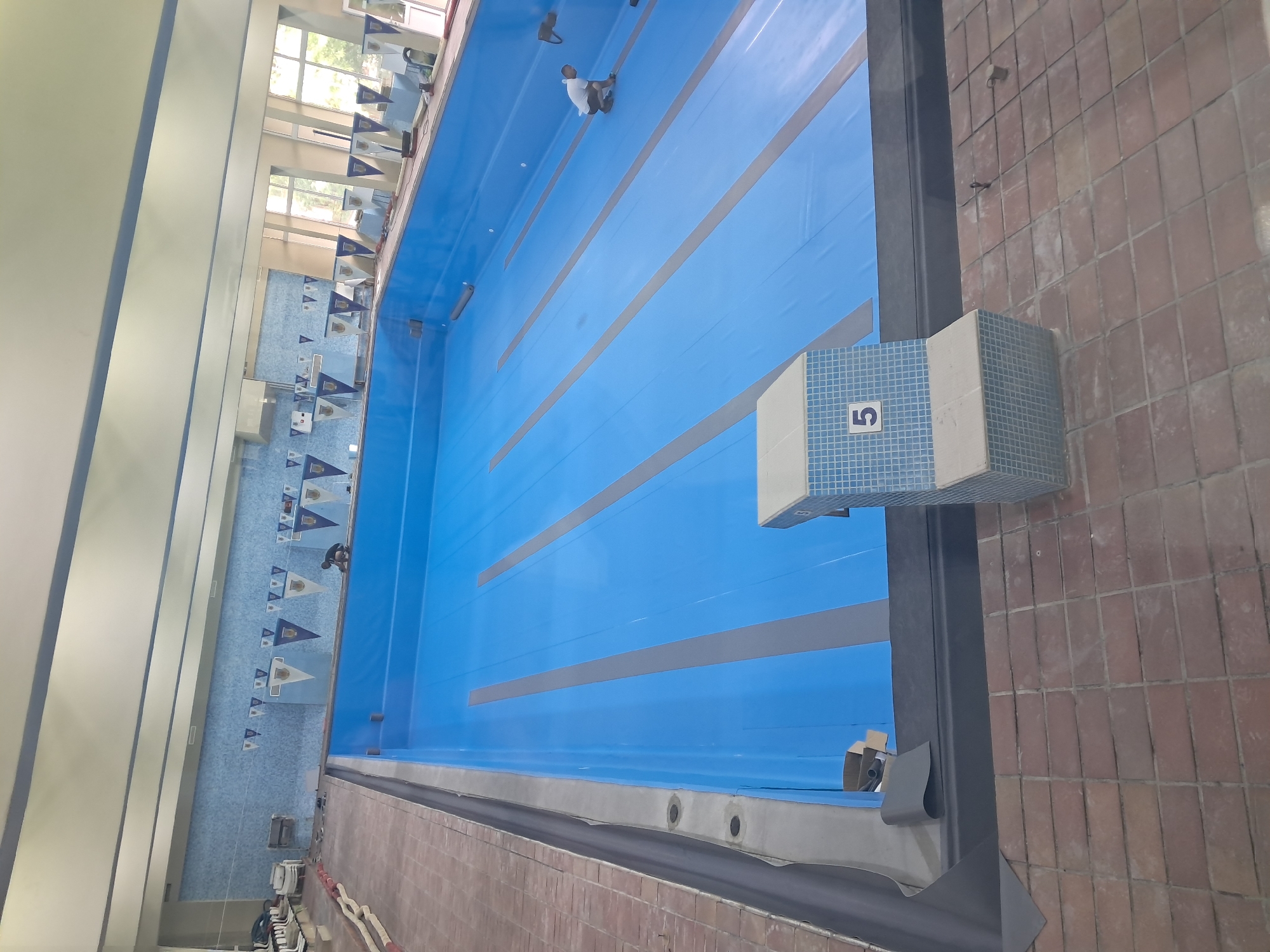 A high-angle shot depicts an indoor swimming pool undergoing renovation in Veliko Tarnovo, Bulgaria. The pool, filled with blue lining, features dark gray lane markers. On the right side of the frame, a starting block with the number "5" is visible. A man in white clothing is seen working on the far side of the pool. Pennant flags hang along the sides of the pool. The pool's surrounding area, including the walls and flooring, appears to be in the process of being updated or repaired. Brown brick flooring is also visible.