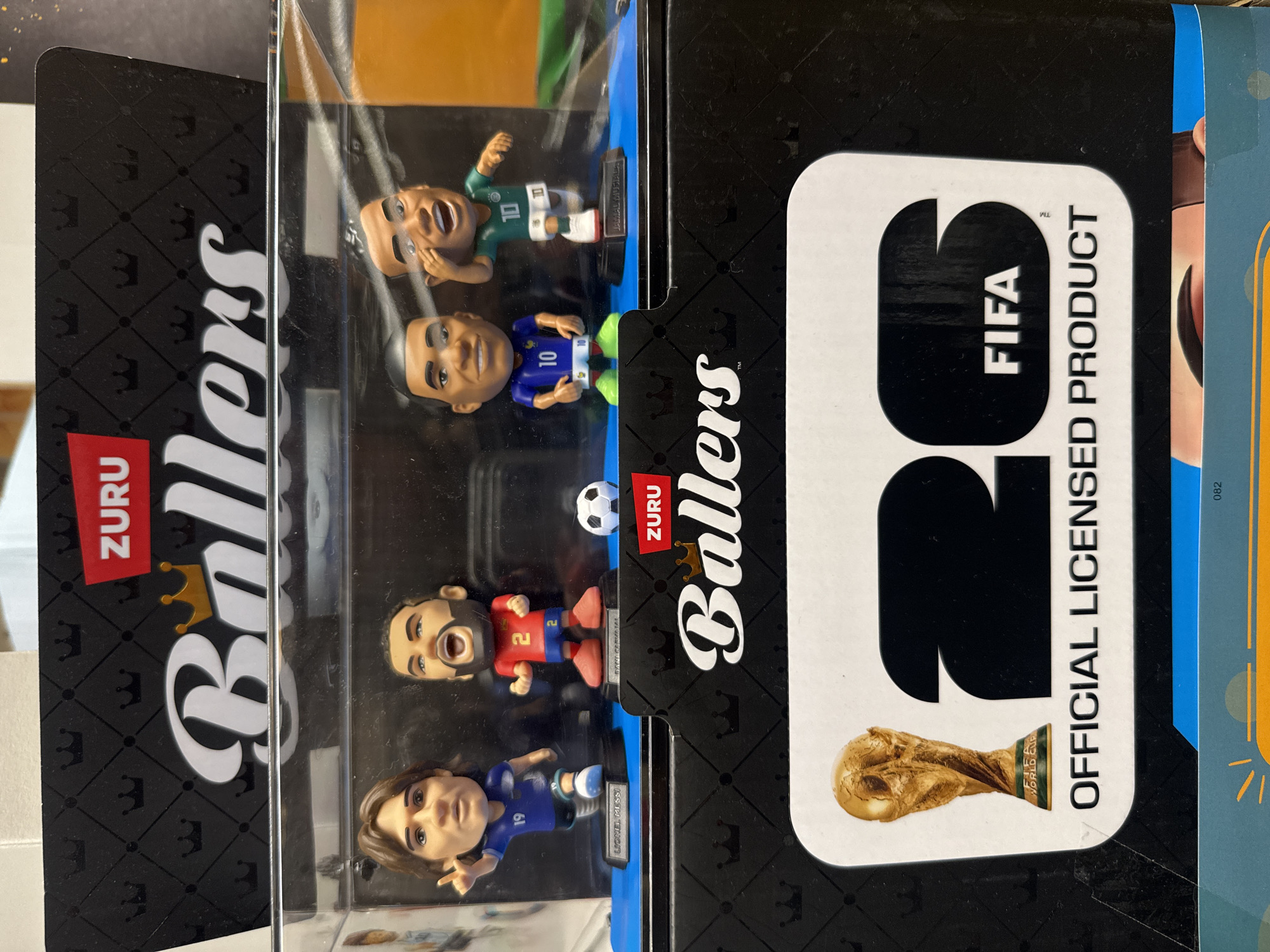 The image shows a product display featuring four ZURU Ballers soccer player figurines inside a clear plastic case. The figurines are cartoonish representations of famous soccer players. The display box has black and white branding with crowns and the "ZURU Ballers" logo. Below the figurines, there is a larger graphic displaying the FIFA World Cup trophy, the number "26", and the text "OFFICIAL LICENSED PRODUCT". The background of the product is black with a repeating crown pattern. The figurines are positioned on blue bases. The setting appears to be a retail environment, likely a store shelf or display.