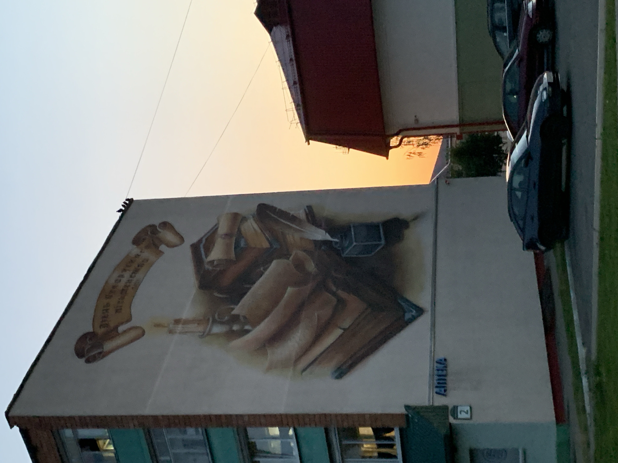 A mural depicting a quill, inkwell, and scrolls is painted on the side of a multi-story building in the city of Kapyĺ, Belarus. The mural is located on the building at street number 2. In the foreground, several cars are parked, and a patch of green grass is visible. The sky above is a gradient of blue and orange, indicating either sunrise or sunset.
