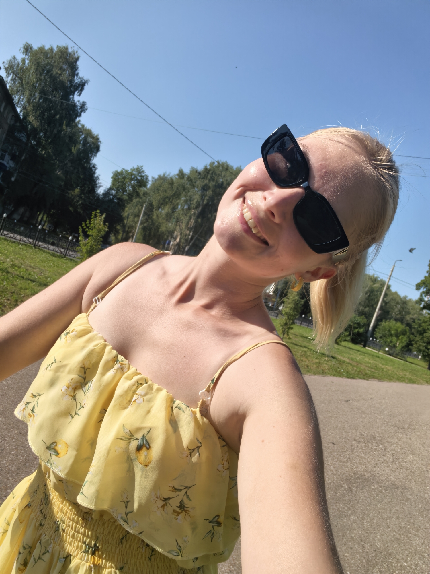 In a low-angle, outdoor, close-up shot, a fair-skinned woman with blonde hair tied in a ponytail smiles broadly while wearing black, square-framed sunglasses. Her head is tilted back, and her eyes are concealed behind the lenses. She is wearing a yellow sundress with a floral and lemon print. The dress features a ruffled neckline and spaghetti straps. Her left arm is visible, bent at the elbow and partially obscuring the lower portion of her face. In the background, lush green trees and a clear blue sky are visible, suggesting a sunny day. The ground is an asphalt path, and a subtle shadow is cast on it. The scene appears to be a park or a tree-lined street in Sterlitamak, Russia.