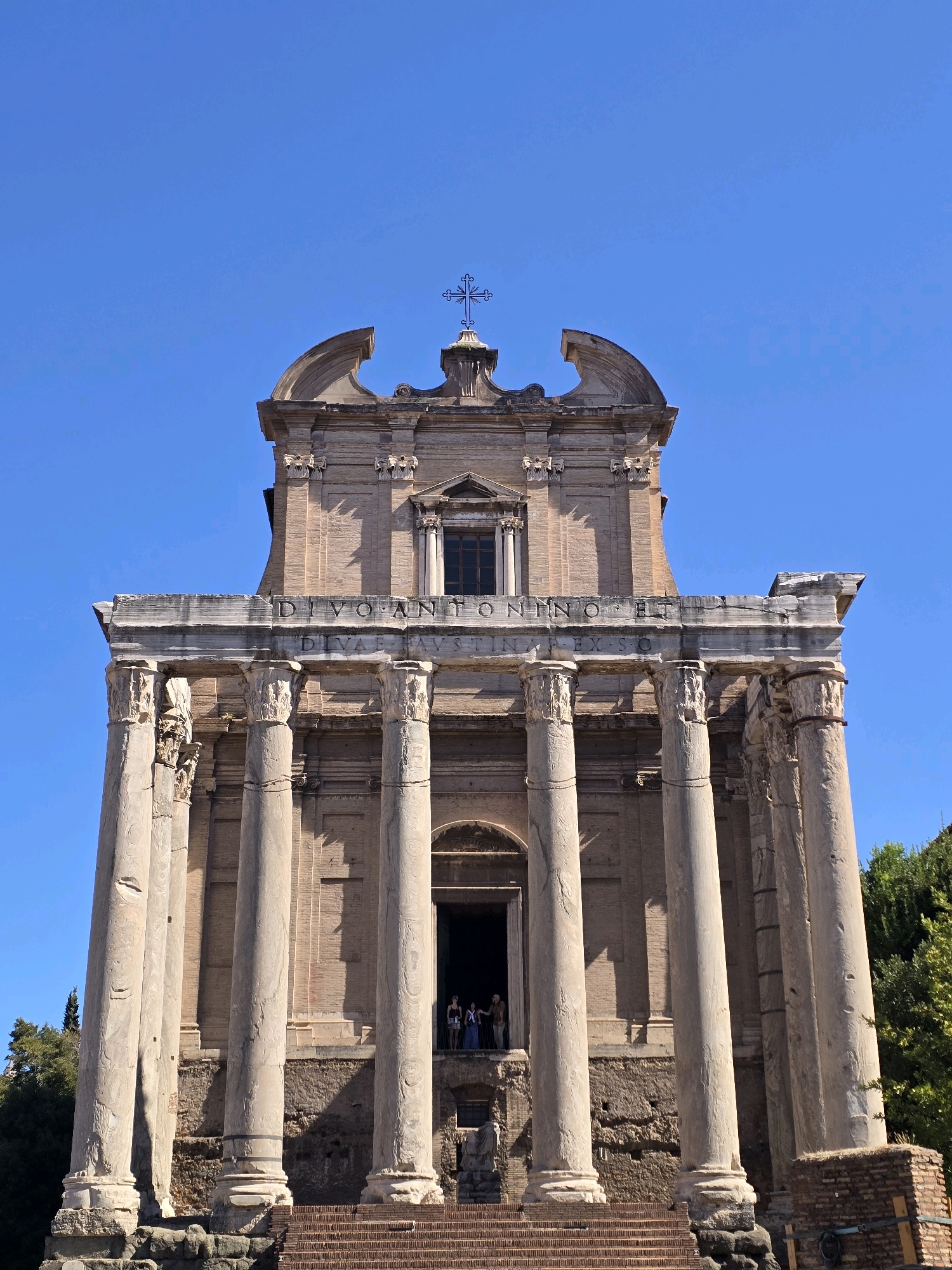 The image shows the Temple of Antoninus and Faustina in Rome, Italy, under a clear blue sky. The temple features a large portico with Corinthian columns. The facade is made of brick and adorned with classical architectural elements. In the distance, a cross is visible on top of the temple. There are people standing in the doorway of the temple, looking out. The surrounding area is partially obscured by trees and foliage. The ground in front of the temple is covered with steps and debris.