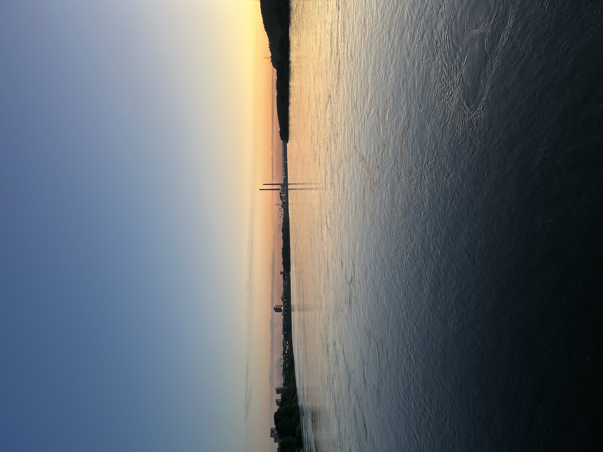 The image shows a body of water reflecting a sky gradient from blue to yellow, suggestive of either sunrise or sunset. On either side of the water, land is visible. The left side shows a city skyline with notable tall structures. At the center of the skyline, a pair of red towers are prominent. Along the right side, there's a strip of land with trees. The water surface displays gentle ripples.