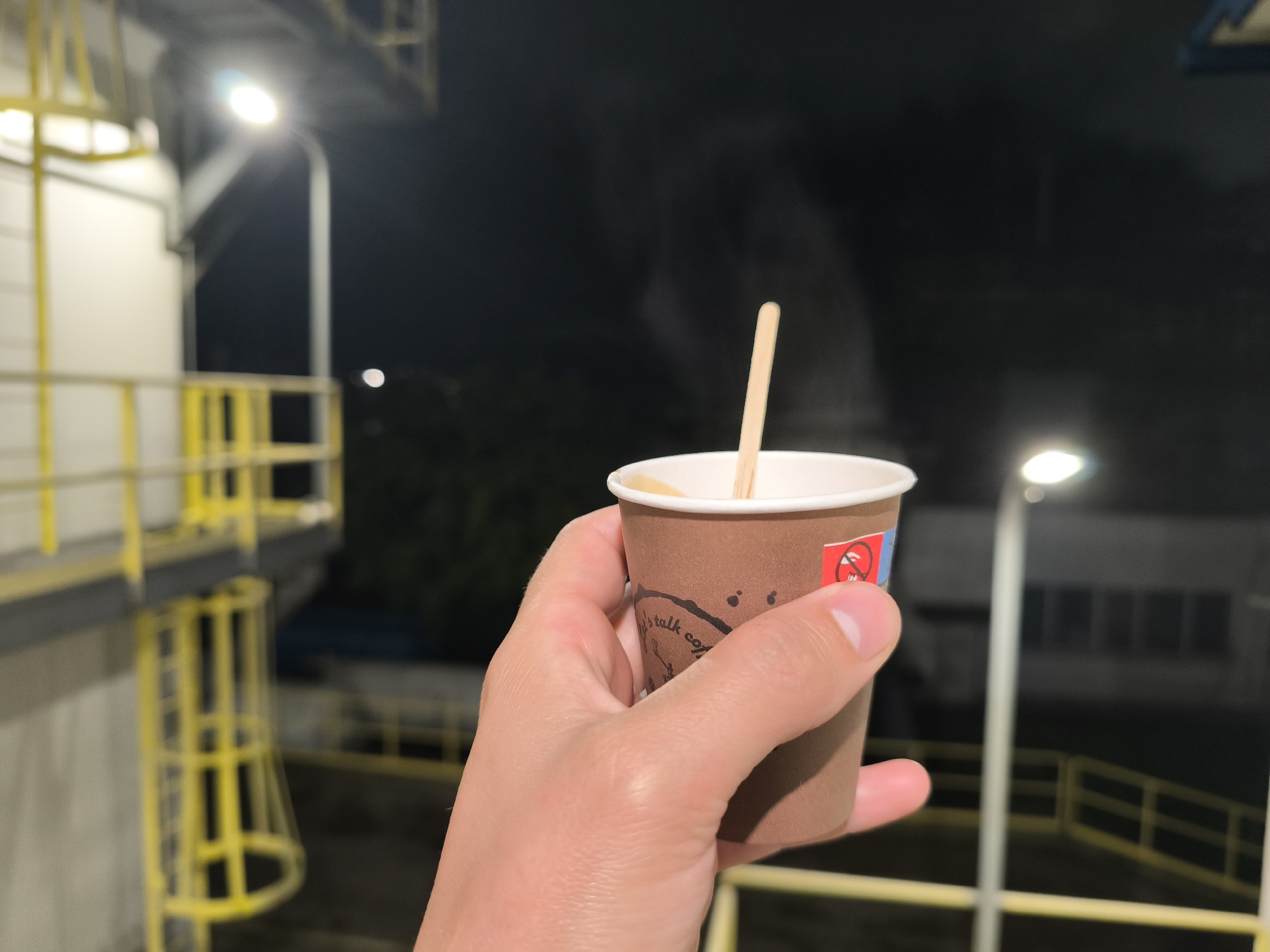 In the foreground, a person's hand holds a brown paper coffee cup with a wooden stirrer. The cup has text and a red and blue sticker with a symbol of a cigarette crossed out. Steam is rising from the cup. The background shows a dark outdoor environment with industrial structures, including a yellow metal staircase and railings, and a bright light fixture. The image appears to have been taken at night.