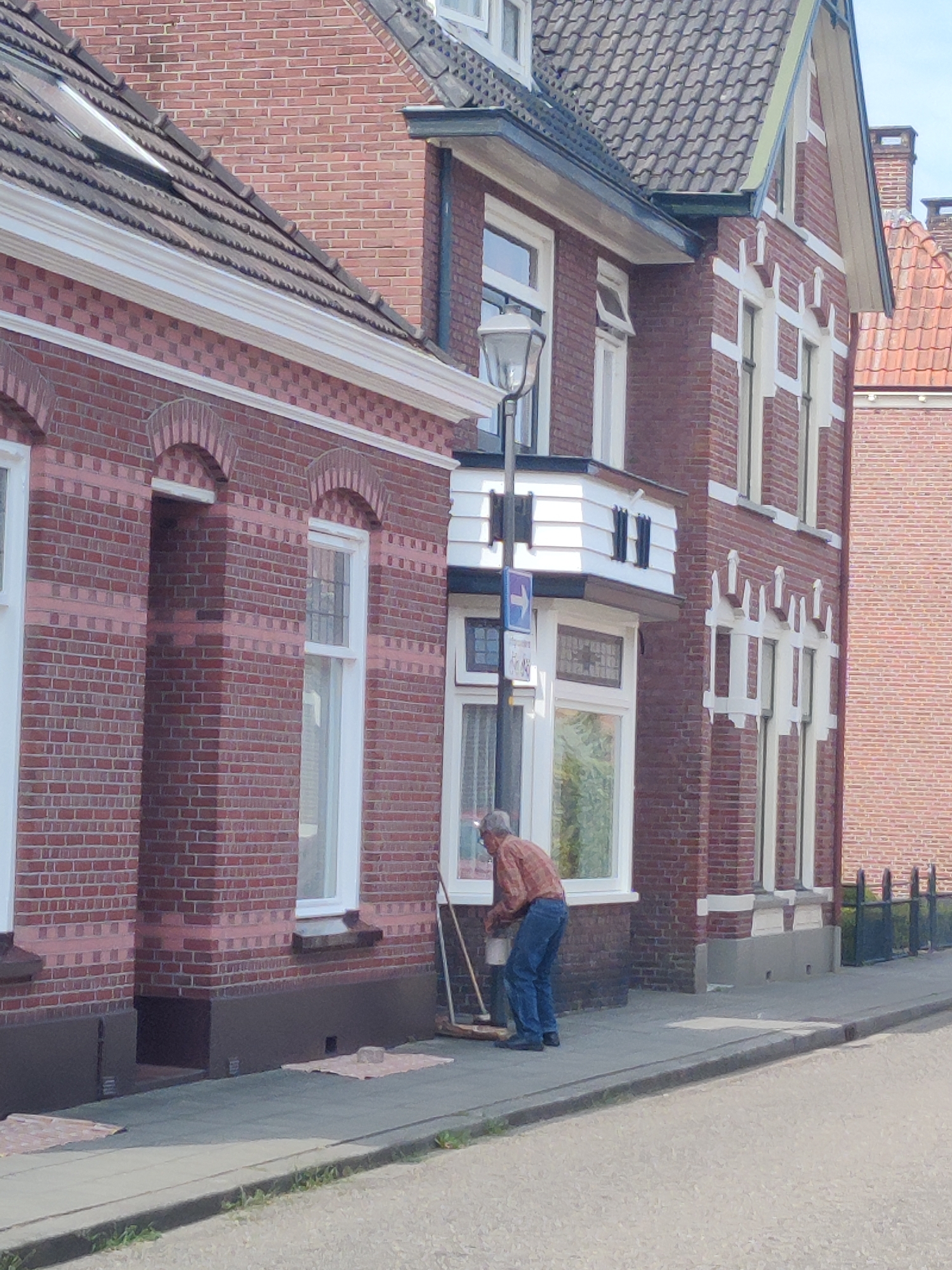 An elderly man, wearing a plaid shirt and blue jeans, is sweeping the sidewalk in front of a red brick building. He is holding a broom and a bucket, and appears to be cleaning the area. The building has multiple windows and decorative white trim. A street lamp is visible on the left side of the image. The scene is set on a sunny day, with the sidewalk and a portion of the street visible in the foreground. The background shows more red brick buildings and some greenery. This scene is located in Winterswijk, Netherlands.