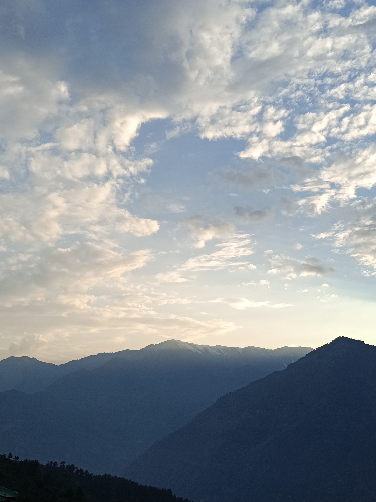 This is a photograph of a mountainous landscape with a dramatic sky. The foreground shows the dark silhouettes of several mountain ranges, with the closest one on the right displaying a distinct, sharp peak dotted with trees. The middle ground features more mountain ranges, also in silhouette, with some showing hints of snow-capped peaks. The background is dominated by a vast sky filled with a mix of scattered white clouds and patches of clear blue. The sunlight appears to be breaking through the clouds, casting a soft, warm glow, particularly towards the horizon. The overall impression is one of a serene and majestic natural environment. The location is identified as Damadi, India.