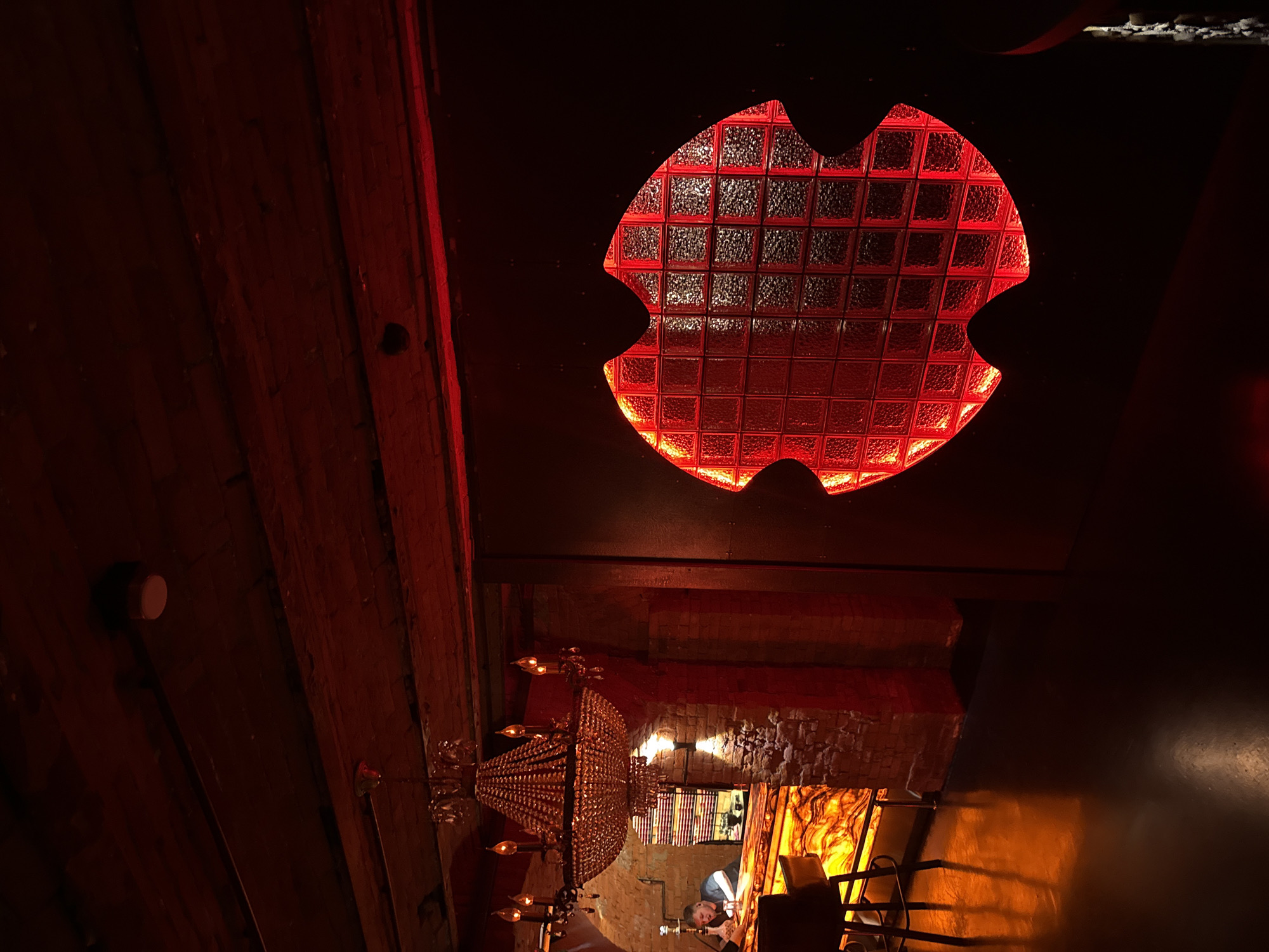This is a dimly lit, high-angle interior shot of a room with exposed wooden beams on the ceiling and a brick wall in the background. The dominant color is red, cast by a large, red-lit window made of glass blocks in an unusual, scalloped shape, possibly resembling a bat or a stylized leaf. Below this window, a large, ornate crystal chandelier hangs, with multiple bulbs emitting a warm light. Further down, a person is partially visible, leaning over a counter or table, appearing to be working on something. The overall atmosphere is moody and somewhat mysterious, with warm lighting creating shadows and highlights. There are no visible signs of text or specific indications of the time of day or weather. The location is identified as Khamkhi, Russia, which might suggest a bar, restaurant, or a unique private dwelling.