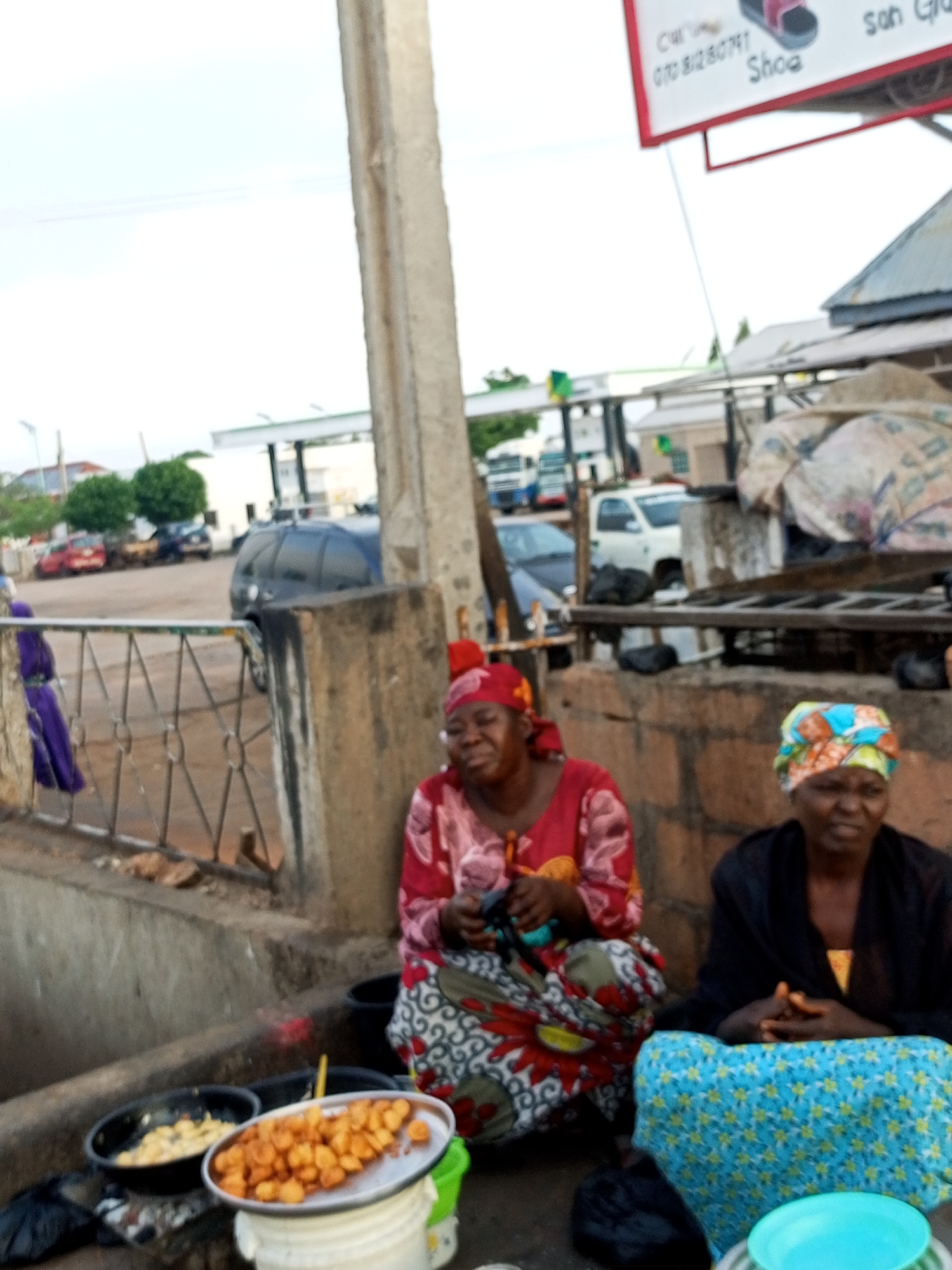 Two women are sitting in front of food stalls near a street in Gombe, Nigeria. The woman on the left is wearing a red headscarf and a patterned dress. She is holding a basket of fried snacks. The woman on the right is wearing a blue and yellow headscarf and a black dress. She is sitting with her hands clasped in front of her. In front of the women are two bowls of food, one containing fried snacks and the other containing a yellow dish. Behind them, there is a fence and several parked cars. In the background, there are some buildings and trees. The sky is overcast.