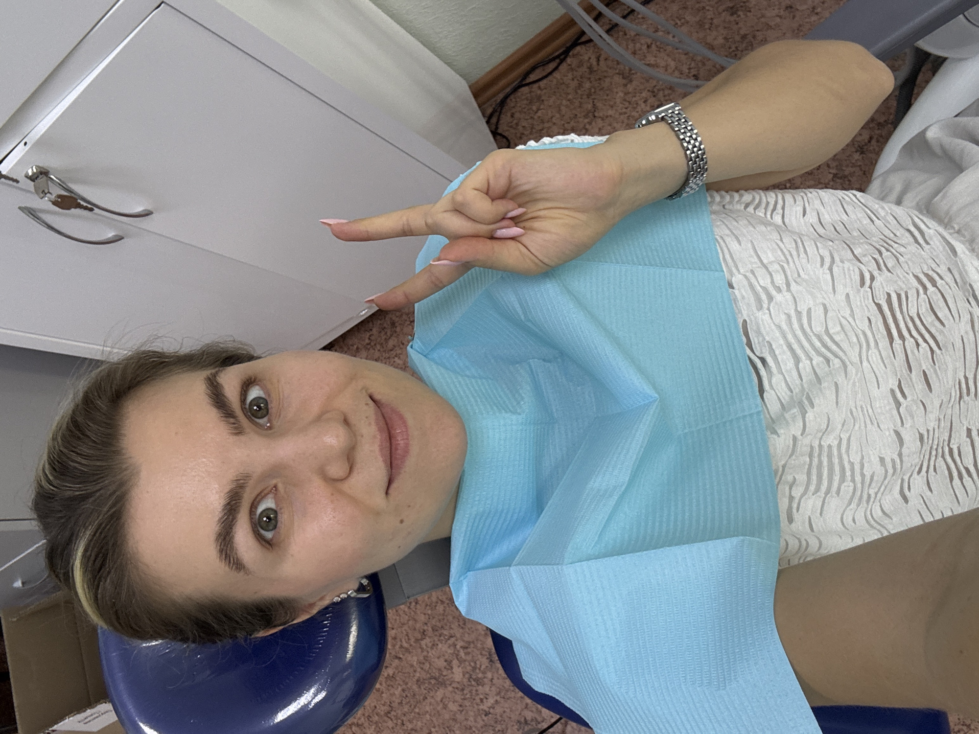 A woman with light brown hair pulled back and blonde highlights is seated in a blue dental chair, wearing a light blue dental bib and a white textured top. She is holding up two fingers in a peace sign gesture with her right hand, which has light pink painted fingernails. Her left ear is adorned with a silver earring. A silver wristwatch is visible on her right wrist. In the background, a white cabinet with silver handles and a key is present, along with a cardboard box and electrical cords. The flooring is a speckled brown. The image appears to be a selfie taken in a dental office.
