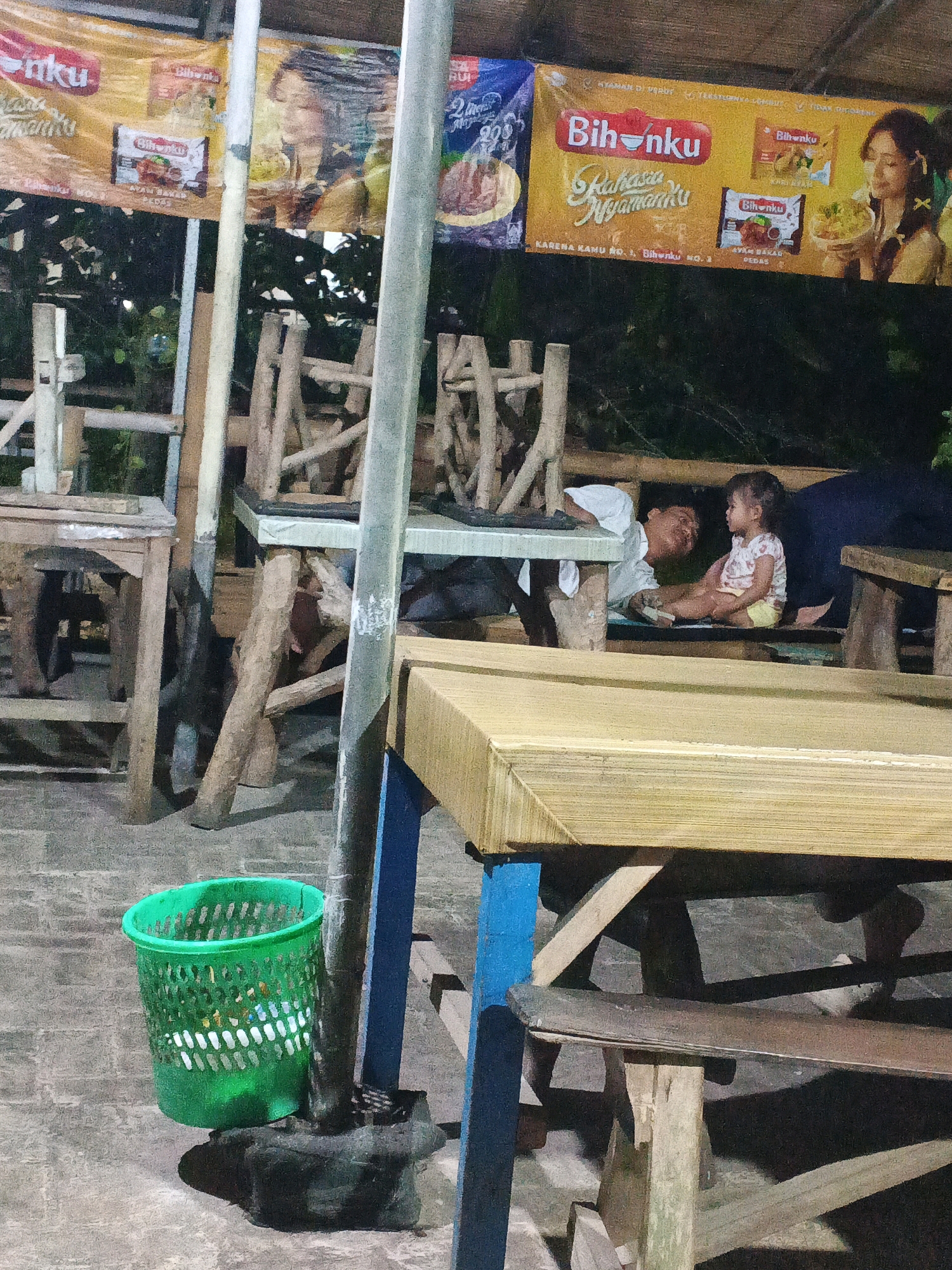 In Ungaran, Indonesia, a father and daughter are seated at a table. The father, wearing a white shirt, leans towards his daughter, who is dressed in a pink and white outfit. They appear to be in a casual outdoor setting, possibly a restaurant or food stall, with wooden furniture and a backdrop of foliage and advertising banners. A green plastic trash can is visible in the foreground, and the overall lighting suggests it is evening or nighttime. The banners advertise "Bihanku," a brand of instant noodles, featuring images of food and smiling individuals.