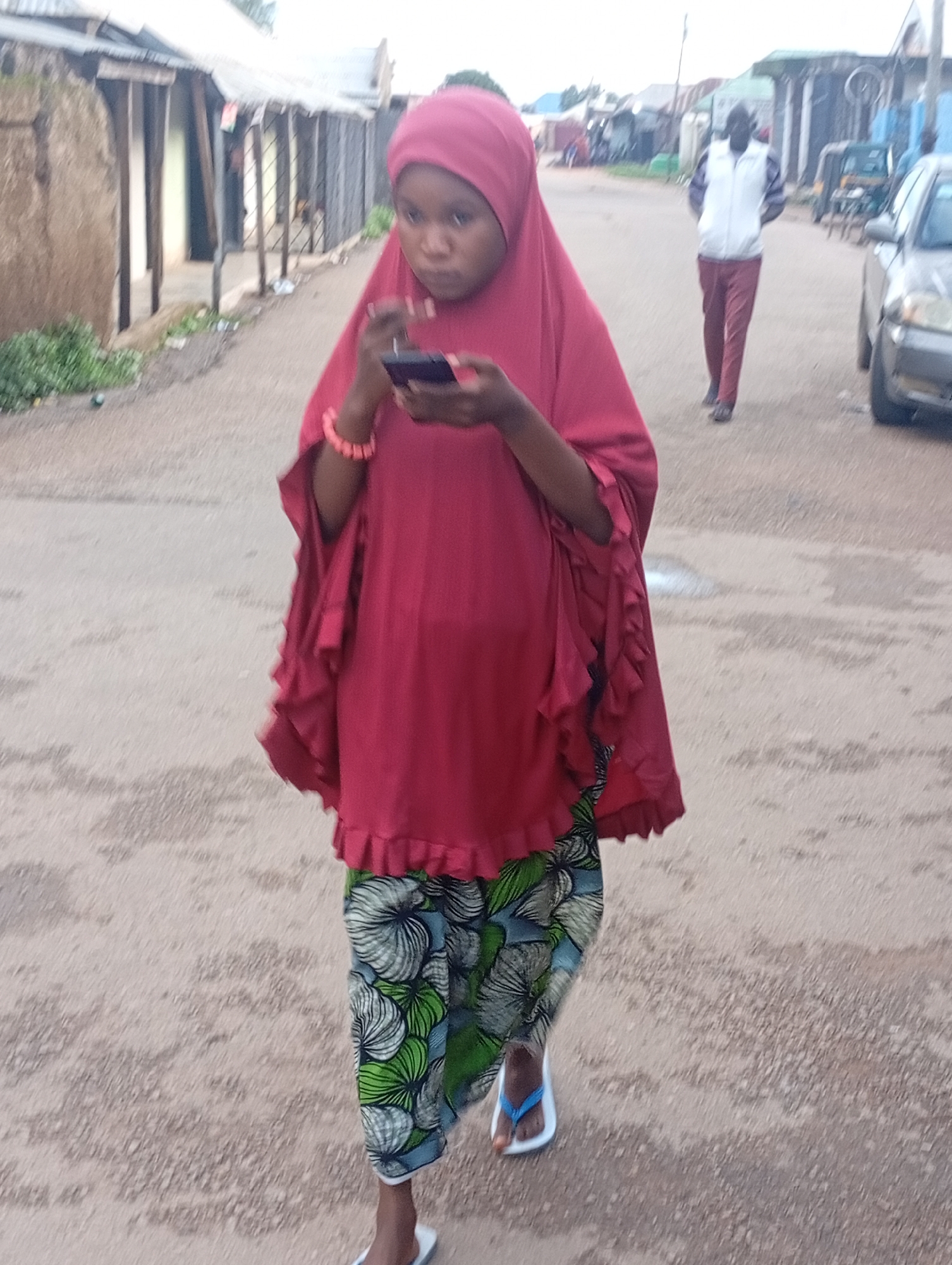 A full-body, medium shot captures a young person wearing a vibrant red hijab and a patterned skirt with white, green, and blue floral designs, walking on a dirt road in an urban environment. The individual is holding a black smartphone and looking down at it, with their left hand adorned with a pink bracelet. A person in the background walks away from the camera, dressed in a white vest over a striped shirt and burgundy pants. The road is lined with simple, weathered buildings, some with corrugated metal roofs and wooden supports. In the distance, more buildings and a hint of greenery are visible, along with a parked car. The ground is a mixture of dirt and gravel, with a few small puddles of water. The lighting suggests it is daytime, with an overcast sky.