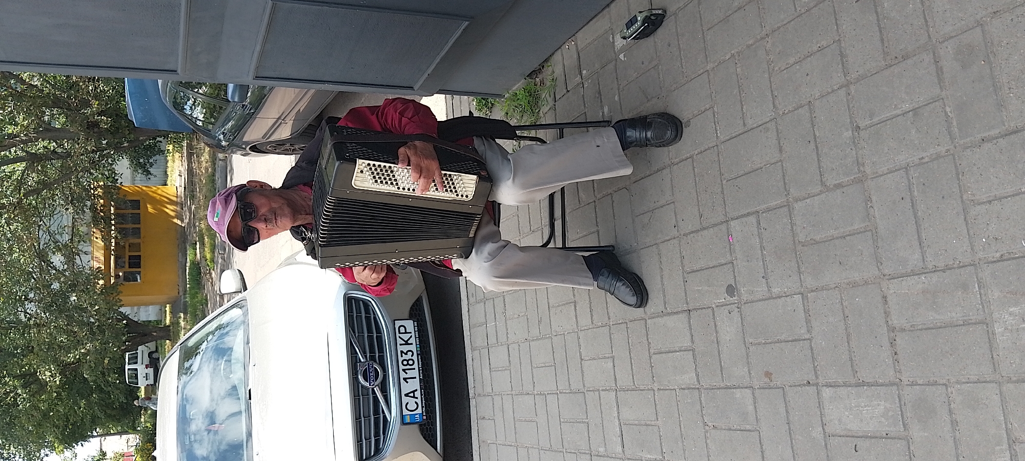 An elderly man wearing sunglasses, a red shirt, a black jacket, and a purple cap is sitting on a chair and playing an accordion. He is positioned in front of a silver Volvo car with a Ukrainian license plate that reads "CA 1183 KP". The scene takes place on a sidewalk paved with gray rectangular bricks. In the background, there is a yellow kiosk and trees, with a white van parked further in the distance. A dark gray modern car is parked to the right of the accordion player, and a dark metal structure is also present on the right side of the image. A small black electronic device is on the ground to the right of the man's feet. The overall setting suggests an outdoor urban environment, likely in Cherkasy, Ukraine.
