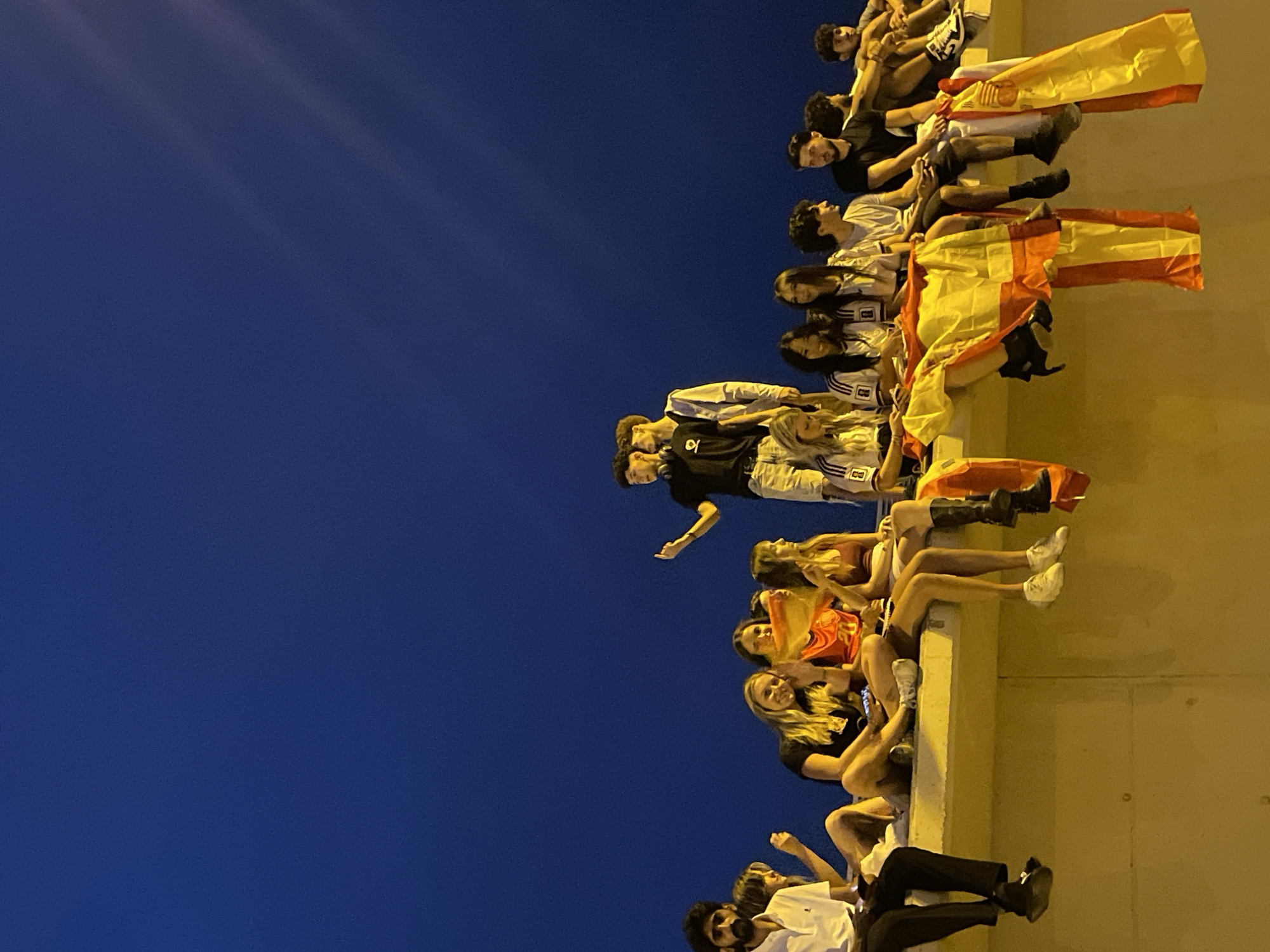 A group of young people, appearing to be fans, are seated on a concrete wall against a dark blue sky. They are dressed casually, with some wearing sports jerseys. Several individuals are holding or draped in Spanish flags. The lighting suggests it is evening or dusk, with artificial light illuminating the wall and the lower portion of the people. The scene takes place outdoors, likely in a public space within Barcelona, Spain, given the prominent display of Spanish flags and the youthful gathering. The individuals are looking in various directions, some interacting with each other and some looking towards the viewer. The overall atmosphere suggests a celebratory or communal gathering.