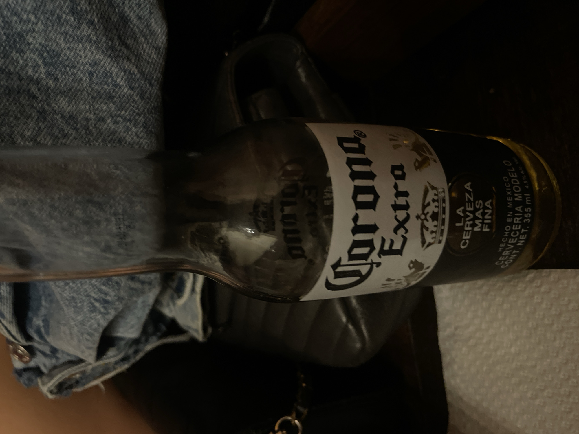 A close-up, medium-shot image displays a Corona Extra beer bottle resting on what appears to be a black handbag or purse, next to a denim jacket or shirt. The angle is slightly tilted, showing the bottle's label prominently. The background is dark, suggesting an indoor setting. No location is indicated in the image.