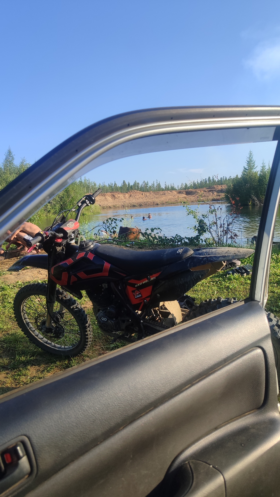 The image depicts a dirt bike parked on grassy terrain next to a body of water, viewed through the open door of a vehicle. The background features a sandy embankment and sparse trees under a clear blue sky. Two individuals are visible in the water, appearing to swim or wade. The dirt bike is black and red with visible branding that reads "OXO MOTORS". The scene appears to be a recreational area, likely a sand or gravel pit converted into a swimming spot. The overall atmosphere is casual and outdoorsy. The location is specified as the city of Mirny, Russia.